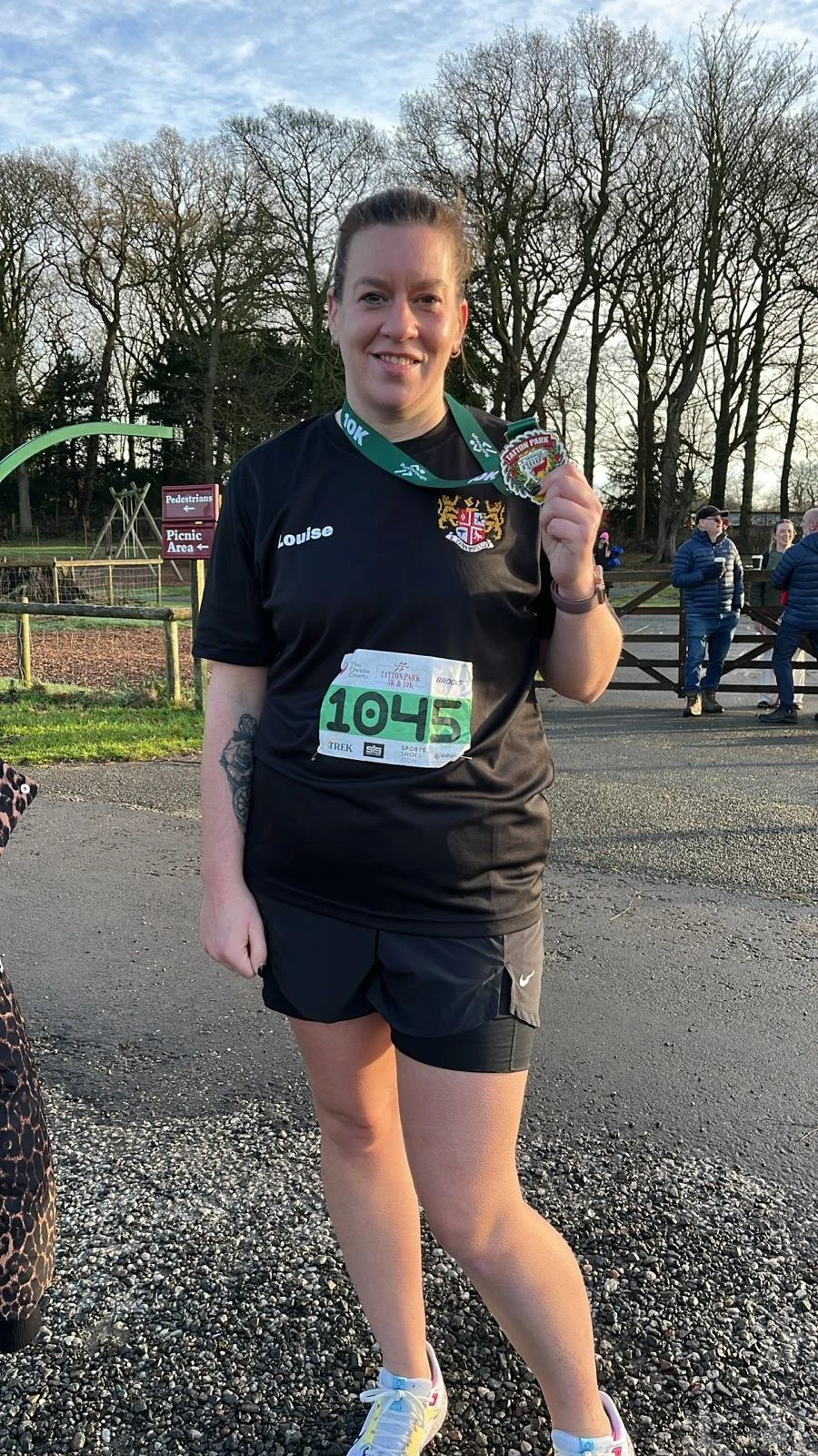 A woman wearing a black race shirt with the name 'Louise' on it, black shorts, and running shoes, standing outdoors after a race, holding a medal, with trees and a few people in the background.
