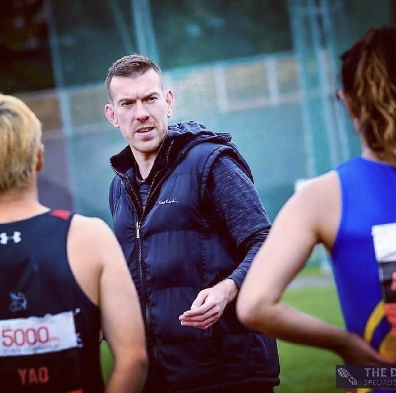 A man with short brown hair and a serious expression, wearing a black vest over a long-sleeve shirt, talking to two female athletes with their backs to the camera on a sports field.