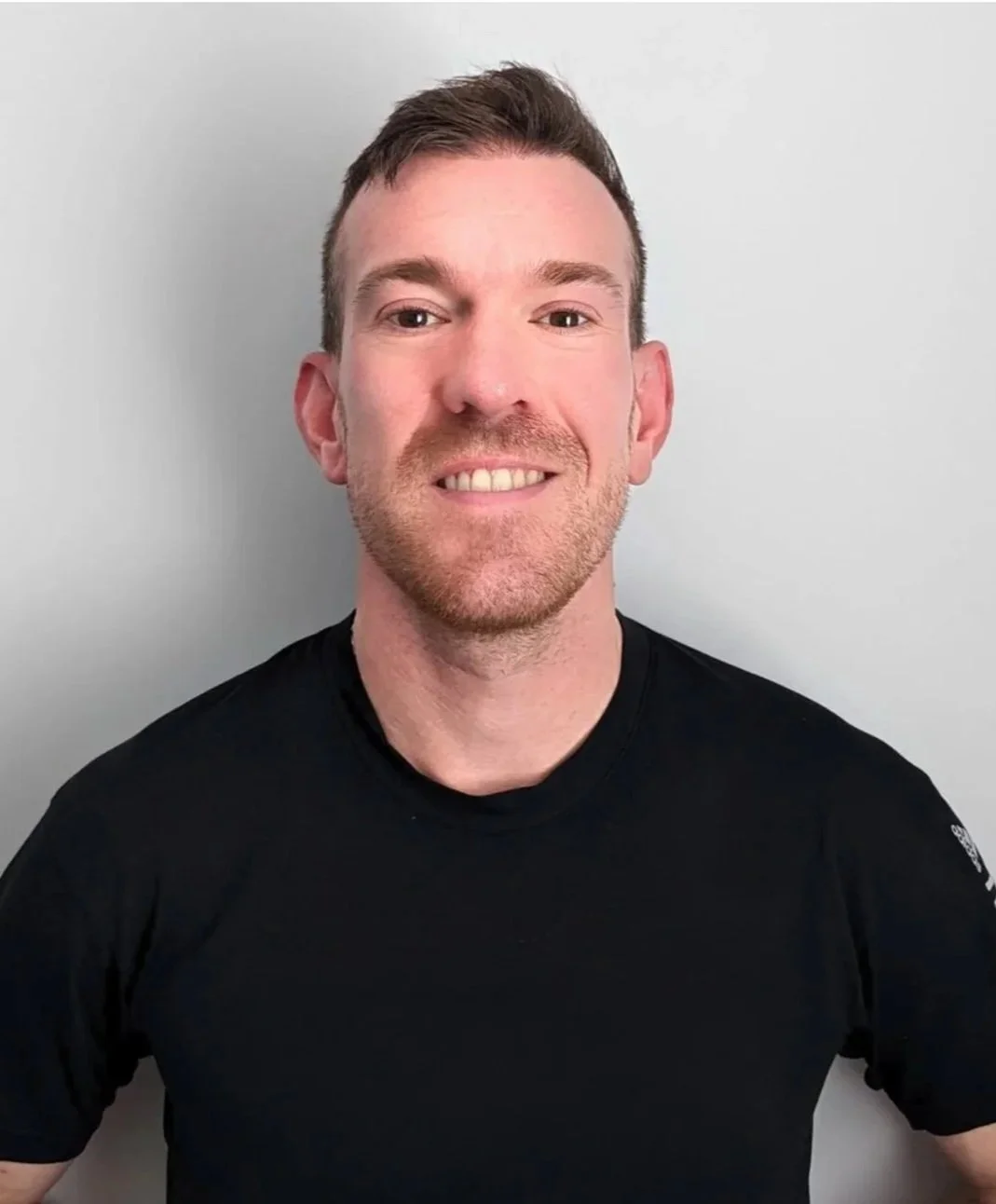 A smiling man with short brown hair, fair skin, and facial hair, wearing a black t-shirt, standing against a light gray wall.