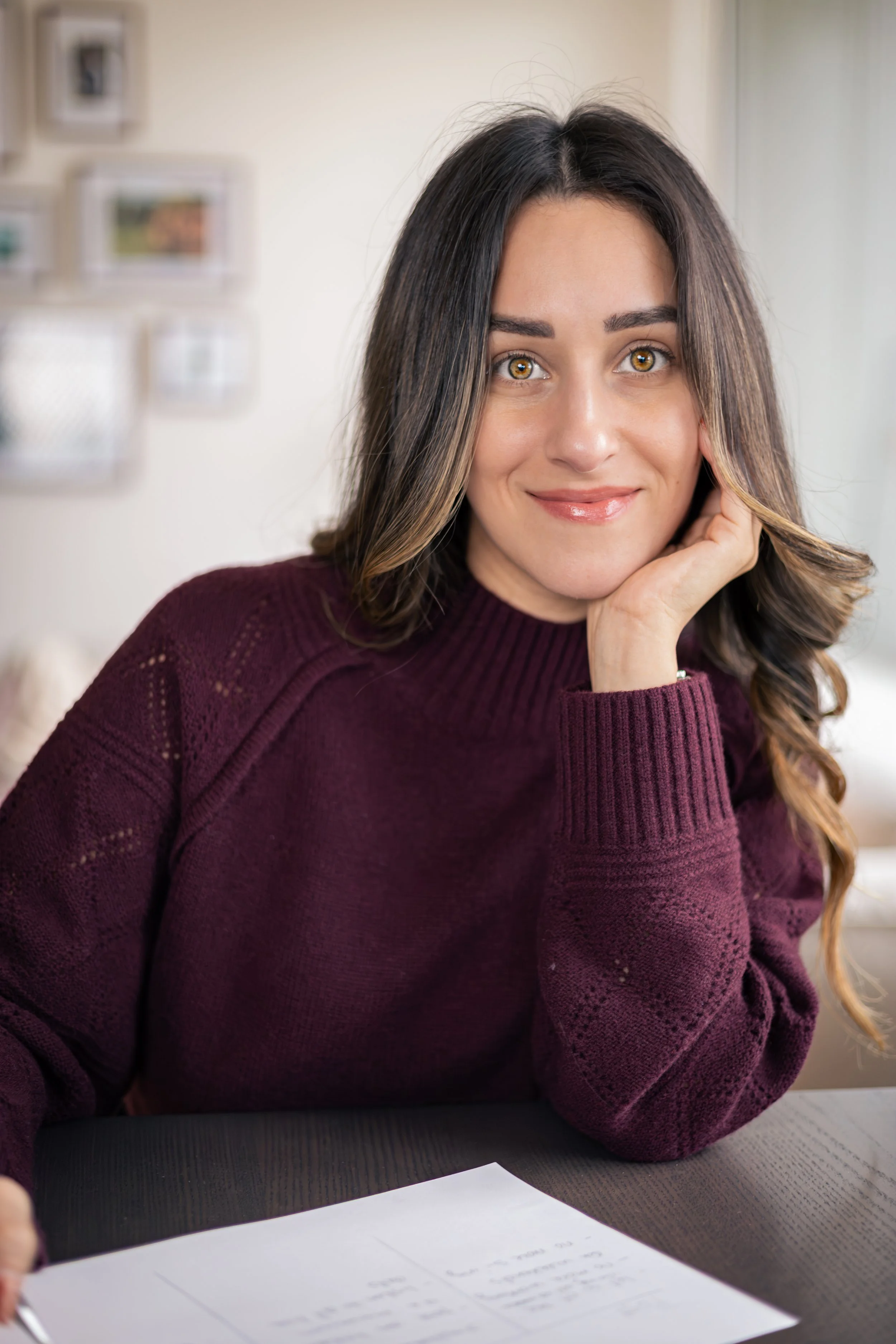 A woman with long brown hair, wearing a maroon sweater, sitting at a table with papers in front of her, resting her face on her left hand and smiling at the camera.