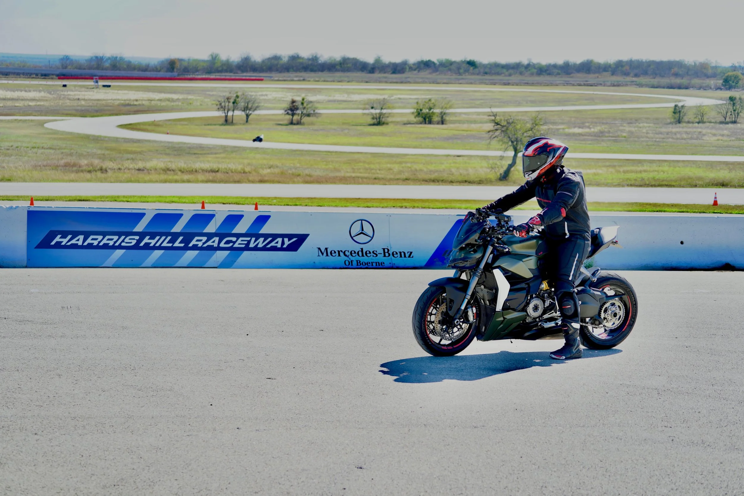 A motorcyclist in full riding gear riding a black and white motorcycle on a race track at Harris Hill Raceway, with a blue and white barrier and grassy landscape in the background.