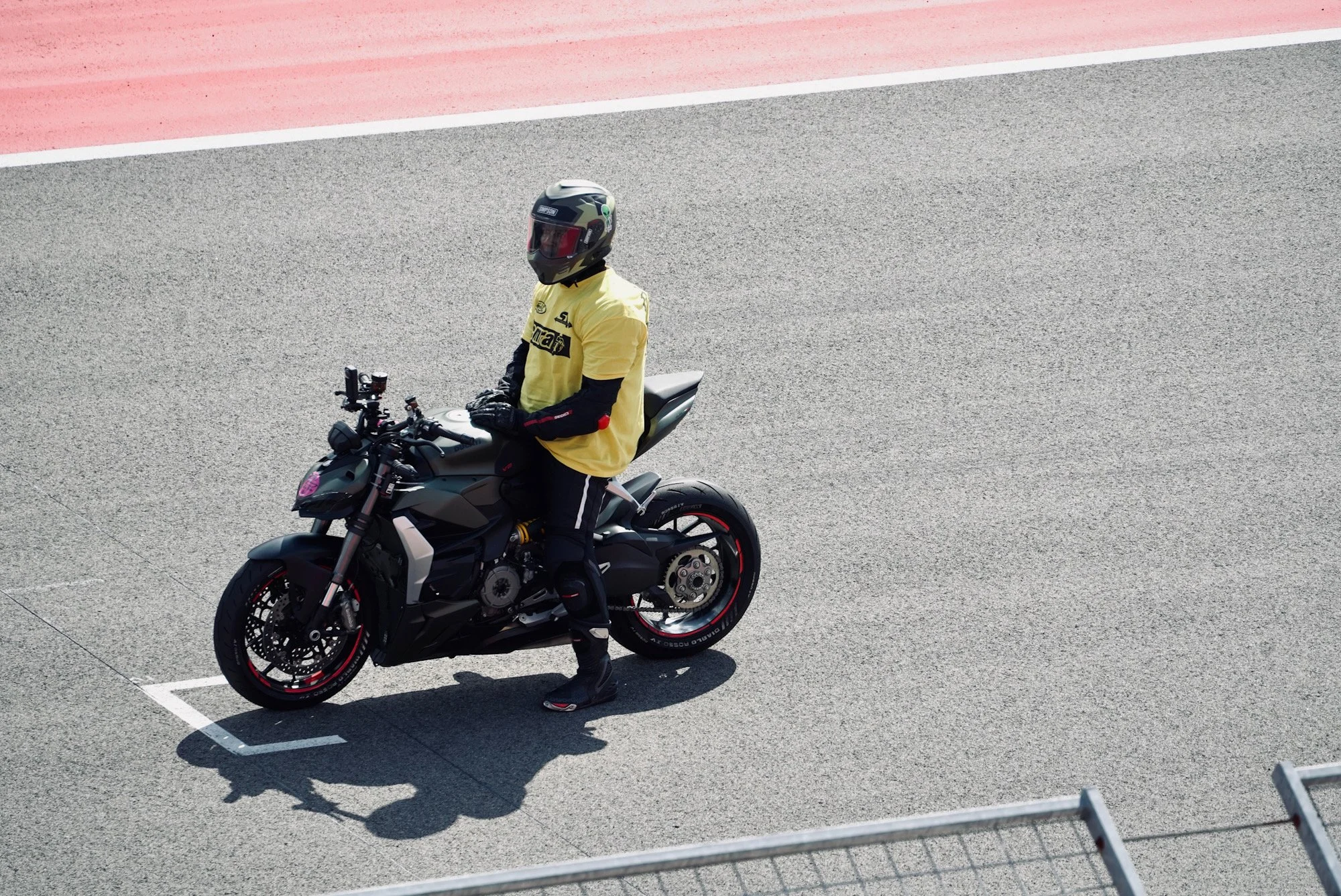 A motorcyclist , black pants, and a full-face helmet sitting on a black motorcycle on a race track with a red and white curb in the background.