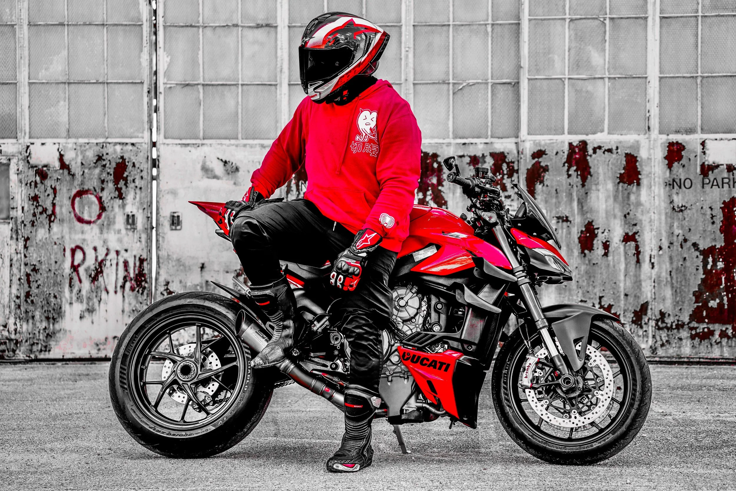 A person wearing a red hoodie with a graphic and Japanese characters, black riding gear, and gloves, sitting on a red Ducati motorcycle in front of a rusted metal gate.