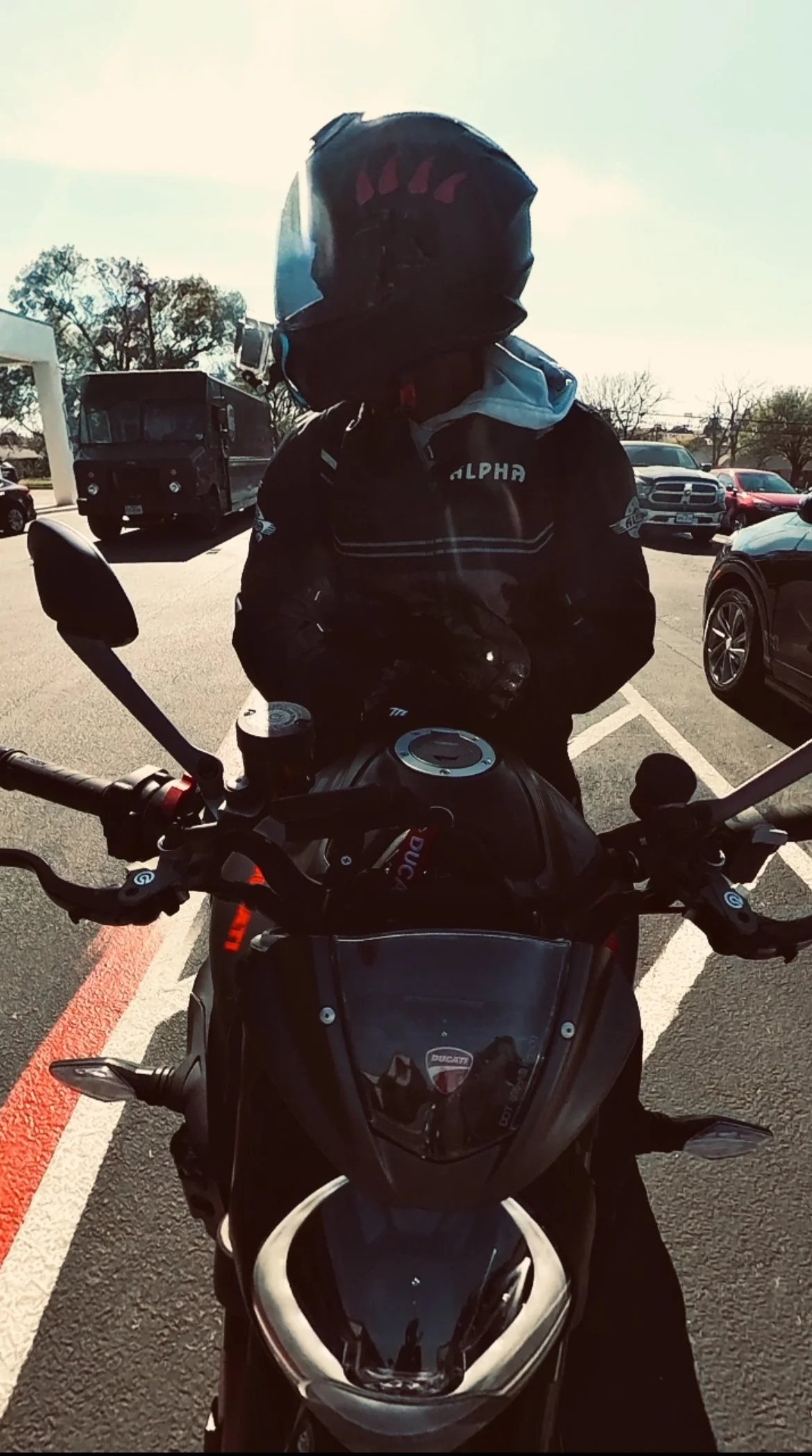 A motorcyclist wearing a black helmet and jacket standing behind a black motorcycle in a parking lot.