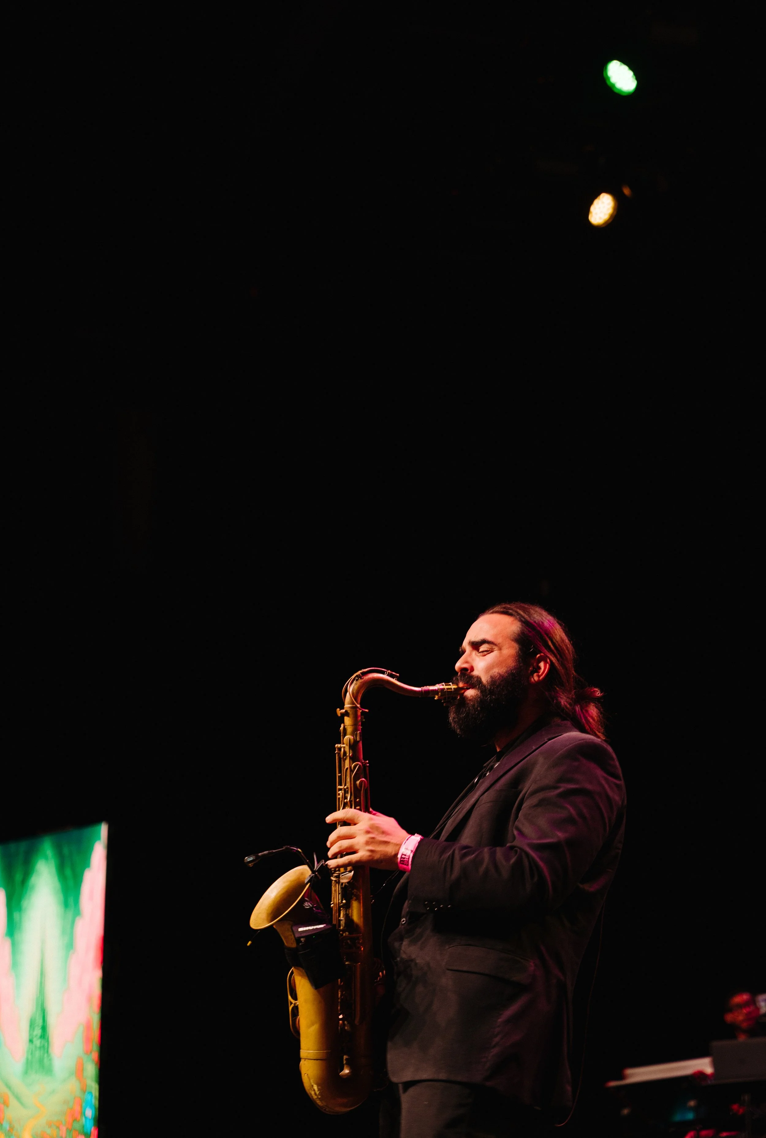 A man with long hair and a beard playing a saxophone on stage with black background and stage lights. Instructor demonstrating musical technique