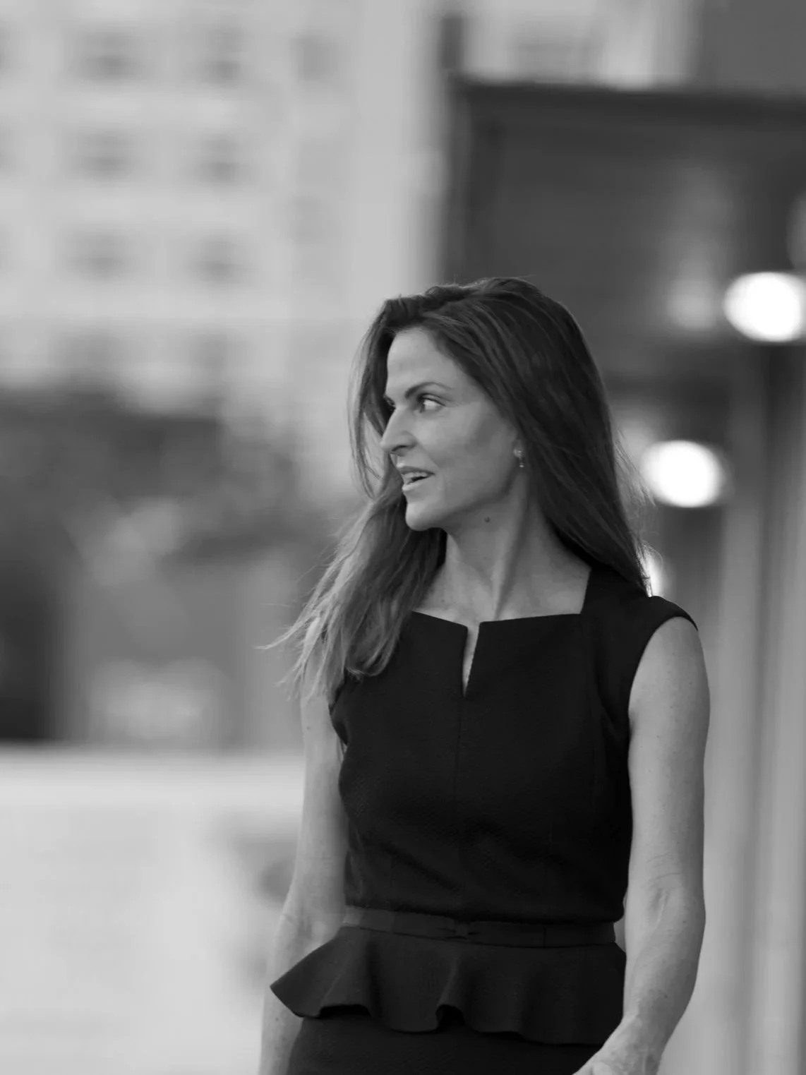 Black and white photo of a woman with long hair, wearing a sleeveless dress with a ruffled waist, looking to her left, standing outdoors with blurred city buildings in the background.