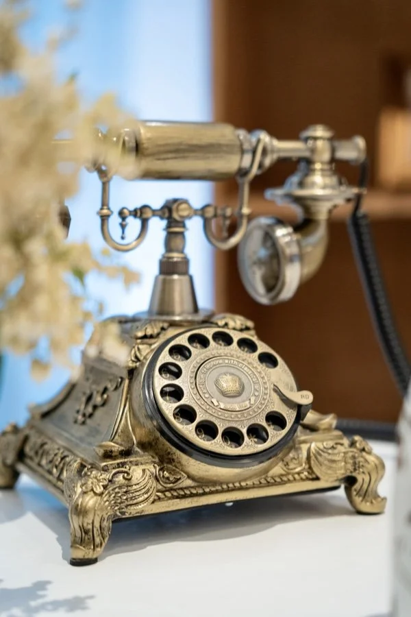 Close-up of an antique rotary telephone with ornate gold-colored details on a white surface, with blurred background.