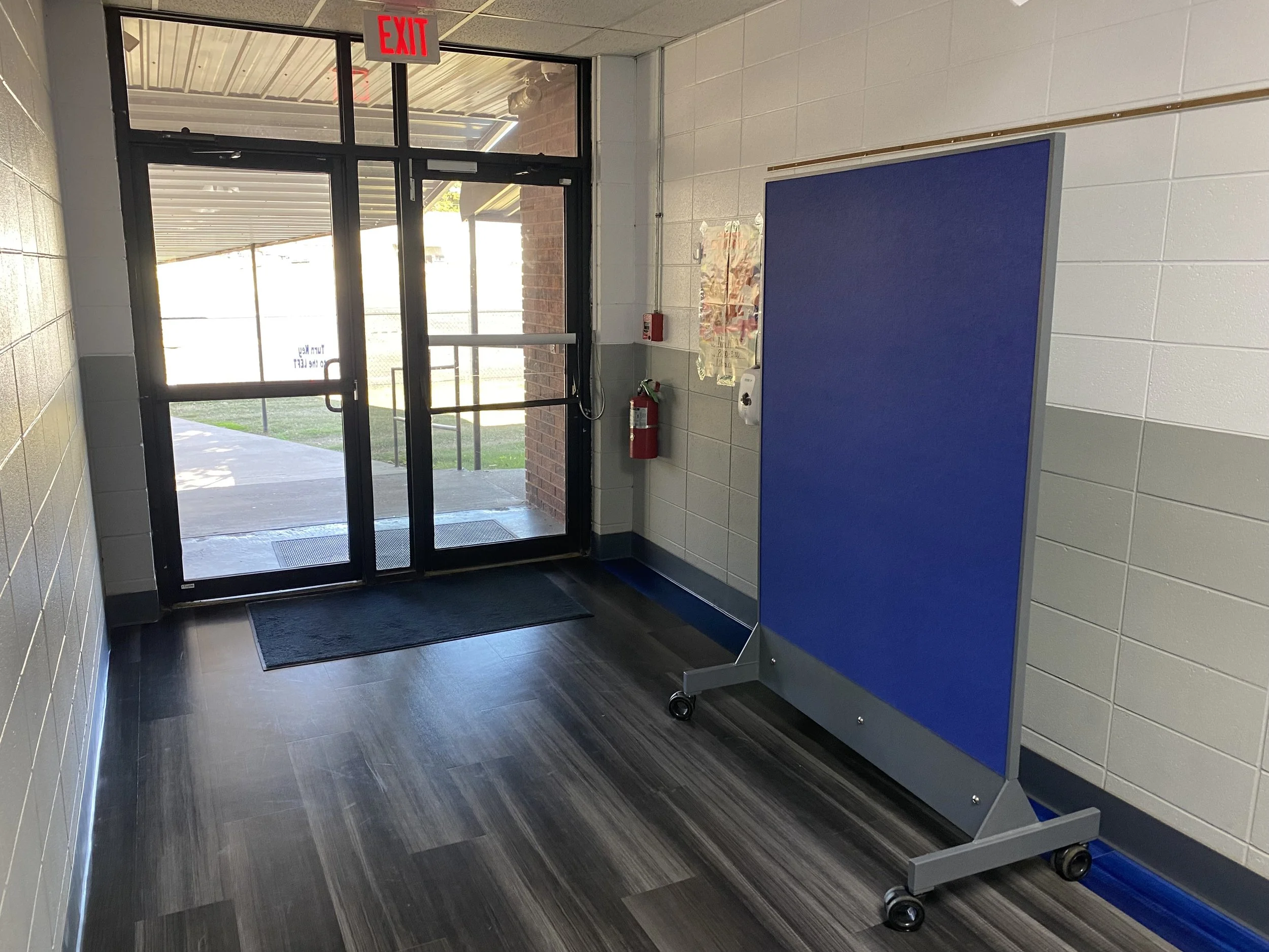 TAG Mobile bulletproof mobile whiteboard with blue acoustic panel positioned next to a school hallway exit door for UL 752 Level 8 ballistic protection.