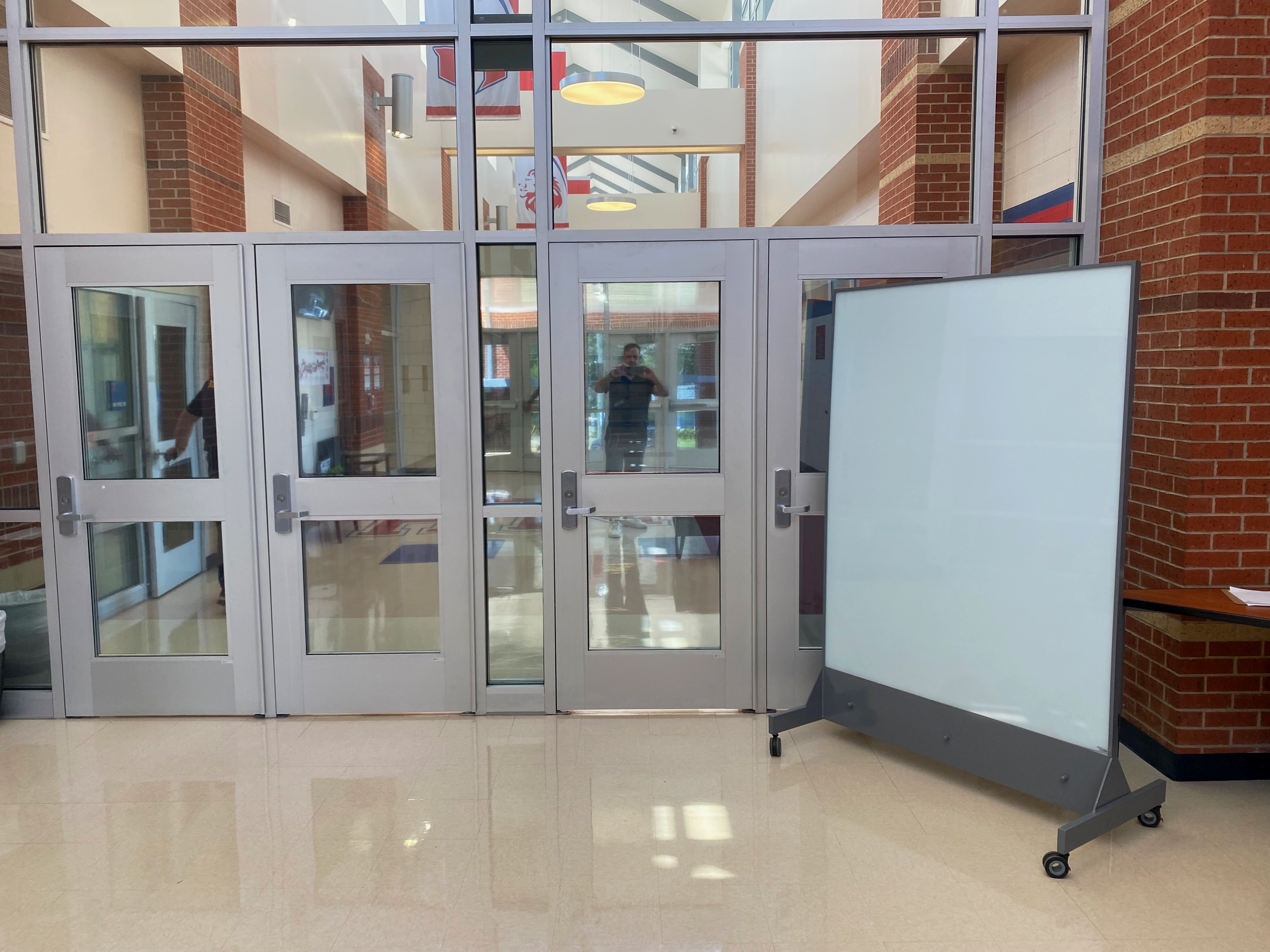 Mobile ballistic glass panel on wheels positioned inside a school entryway near double glass doors, demonstrating use of TAG Mobile unit for added protection.