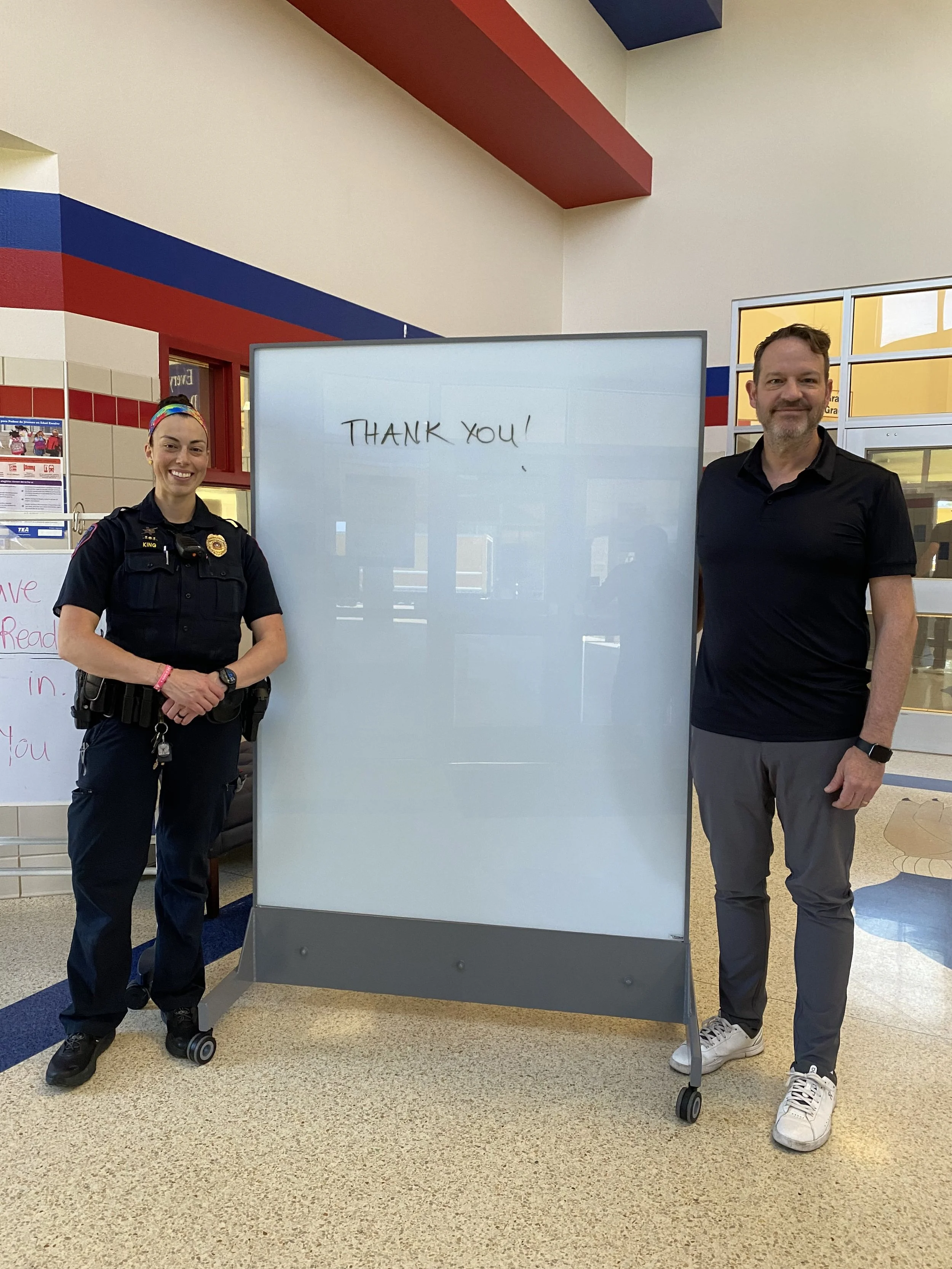 Titan Armored CEO Scott Newman standing with a School Resource Officer in front of a TAG Mobile UL 752 Level 8 bulletproof whiteboard inside a school entryway, featuring “thank you” written on the dry-erase glassboard.