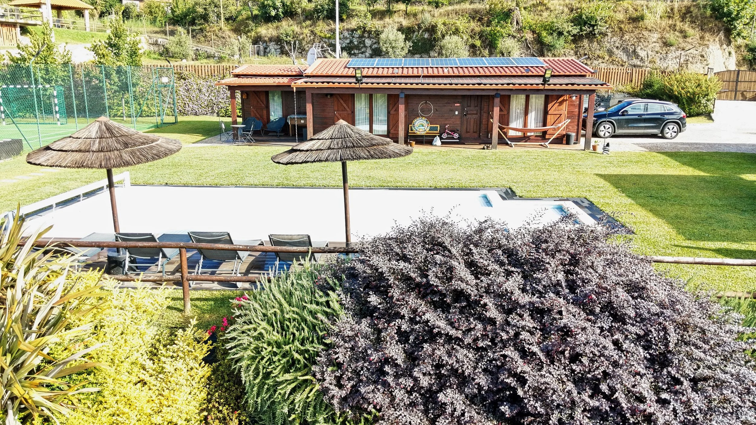 A backyard view featuring a pool with large straw umbrellas, lounge chairs, a grassy yard, bushes, a wooden house with solar panels, a parked car, and a hiking bicycle.