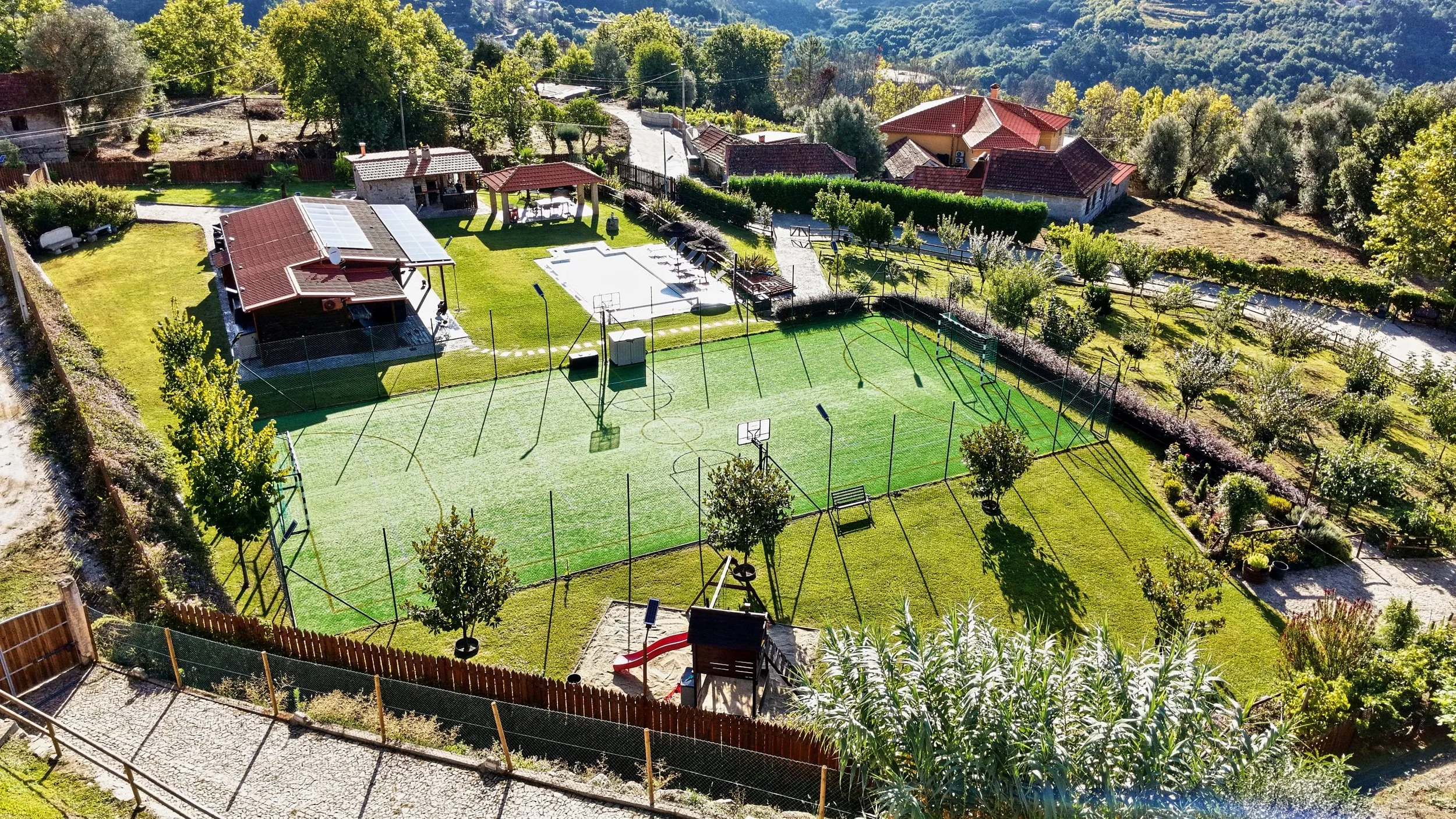 An aerial view of a backyard with a tennis court, a swimming pool, and several small buildings, surrounded by trees and neighboring houses.