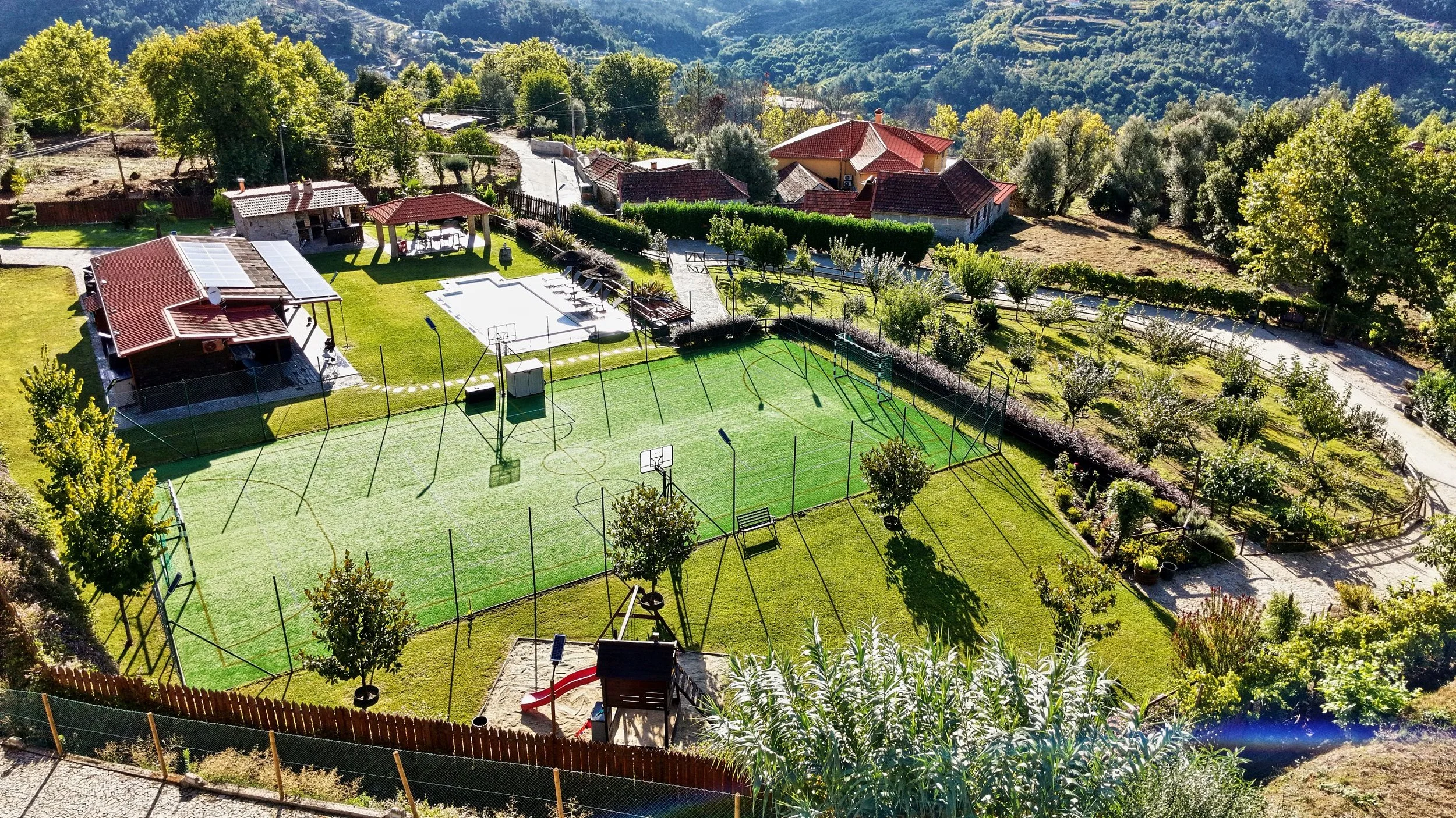 Aerial view of a backyard with a soccer field, basketball courts, a patio with a pergola, a swimming pool, surrounding trees, and nearby houses in a hilly, green landscape.