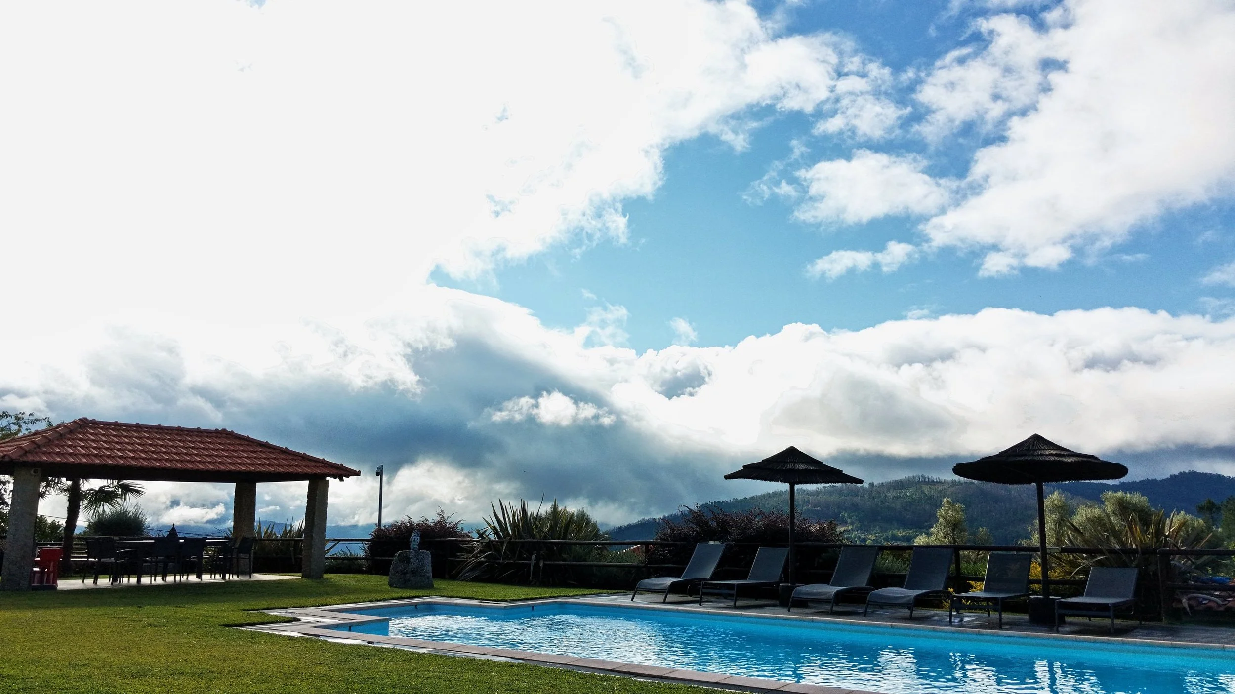 Outdoor swimming pool surrounded by lounge chairs, shaded gazebos, and lush greenery under a partly cloudy sky.