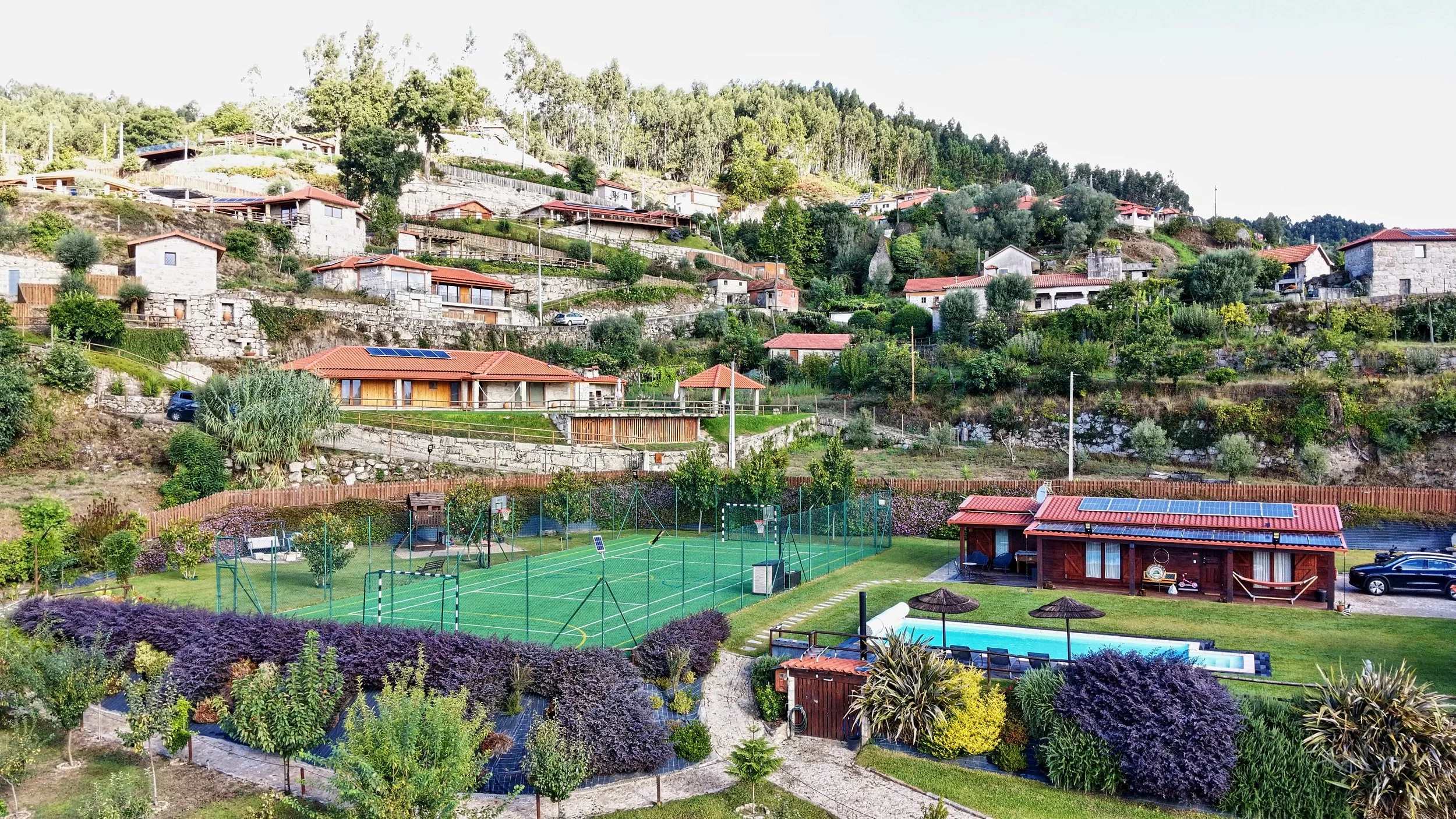 Hilly residential area with houses, gardens, sports courts, a swimming pool, and lush greenery.