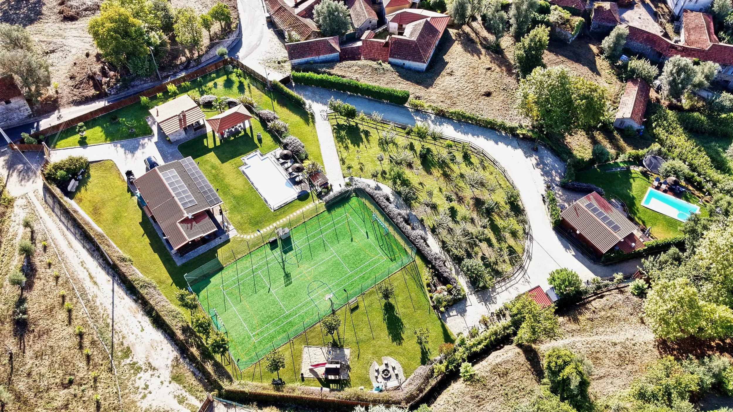 An aerial view of a residential area with houses, a fenced backyard with a small gazebo, a swimming pool, and a sports court, surrounded by trees and dirt paths.