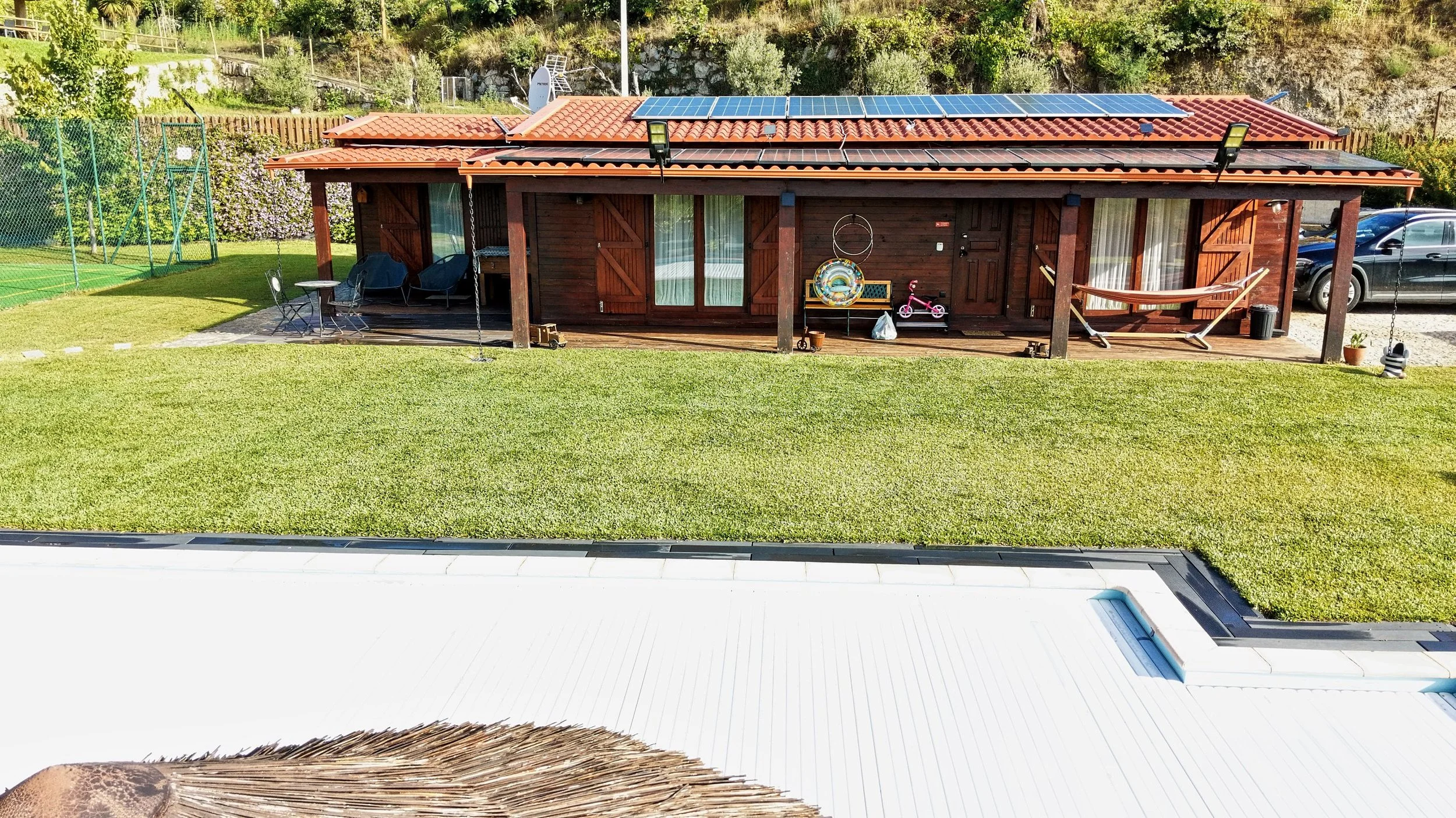 A backyard with a wooden house featuring solar panels on the roof, a grassy lawn, outdoor furniture, children's toys, and a parked black car in the driveway.