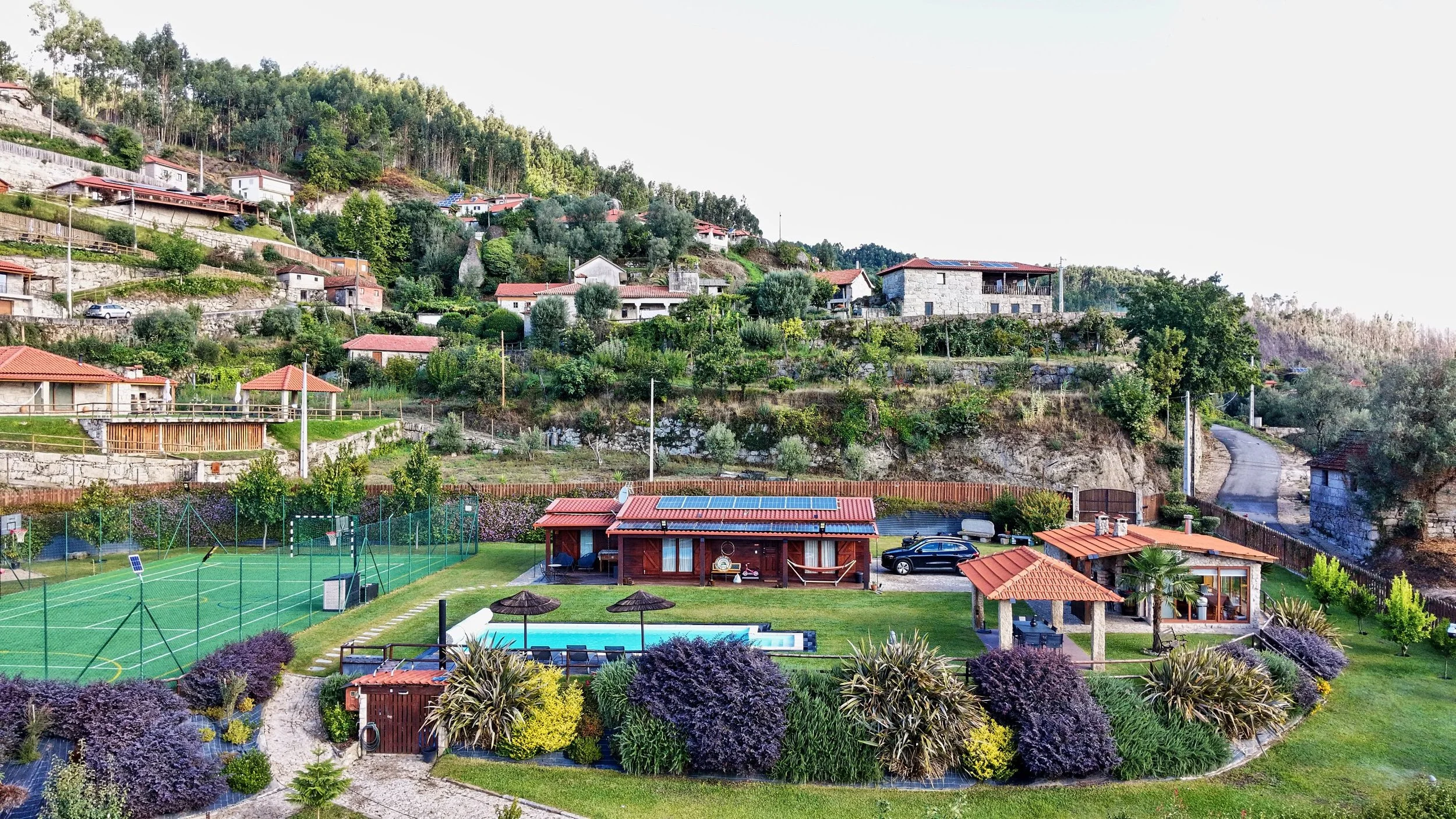 Hilly residential area with houses, lush greenery, and a sports court with tennis or basketball court, a swimming pool, and parked cars.