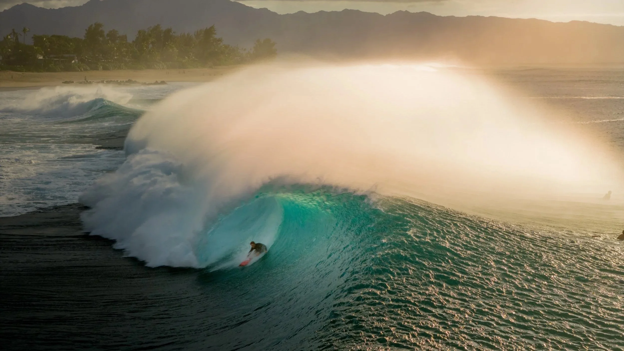 an image of a surfer in a barrel at Pipeline, taken at sunset on the north shore of Oahu. The spray of the wave ignites with sun glow.
