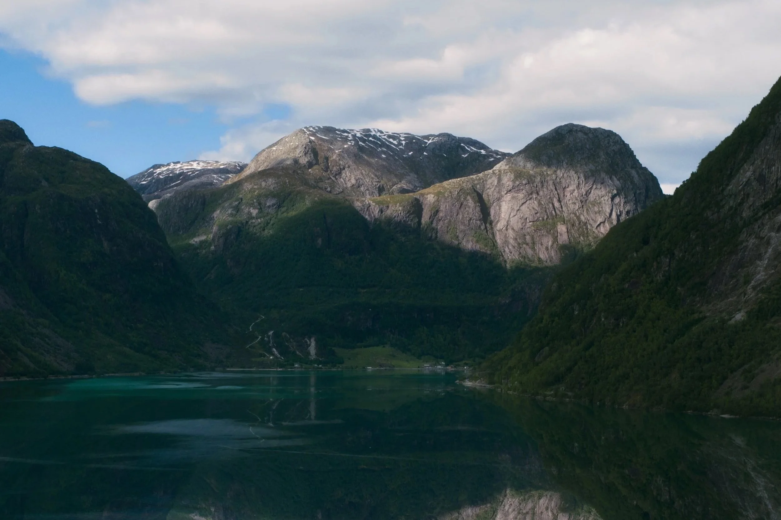 Scenic view of mountainous landscape with rocky peaks, green forested slopes, snow patches on the mountain tops, a calm body of water in the foreground, and a partly cloudy sky.
