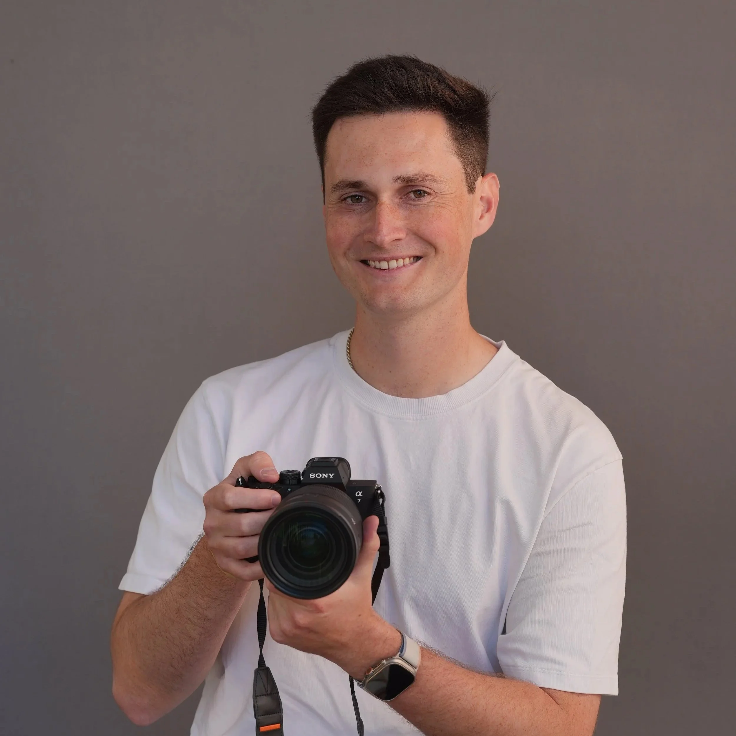 A young man with dark hair and light skin, smiling and holding a camera, wearing a white t-shirt, standing against a plain gray background.