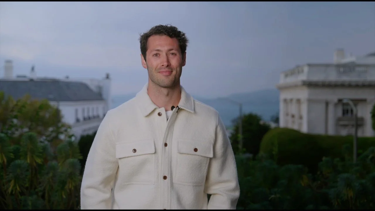 A man stands outdoors in front of a lush landscape with white buildings and mountains in the background, wearing a white jacket and smiling.