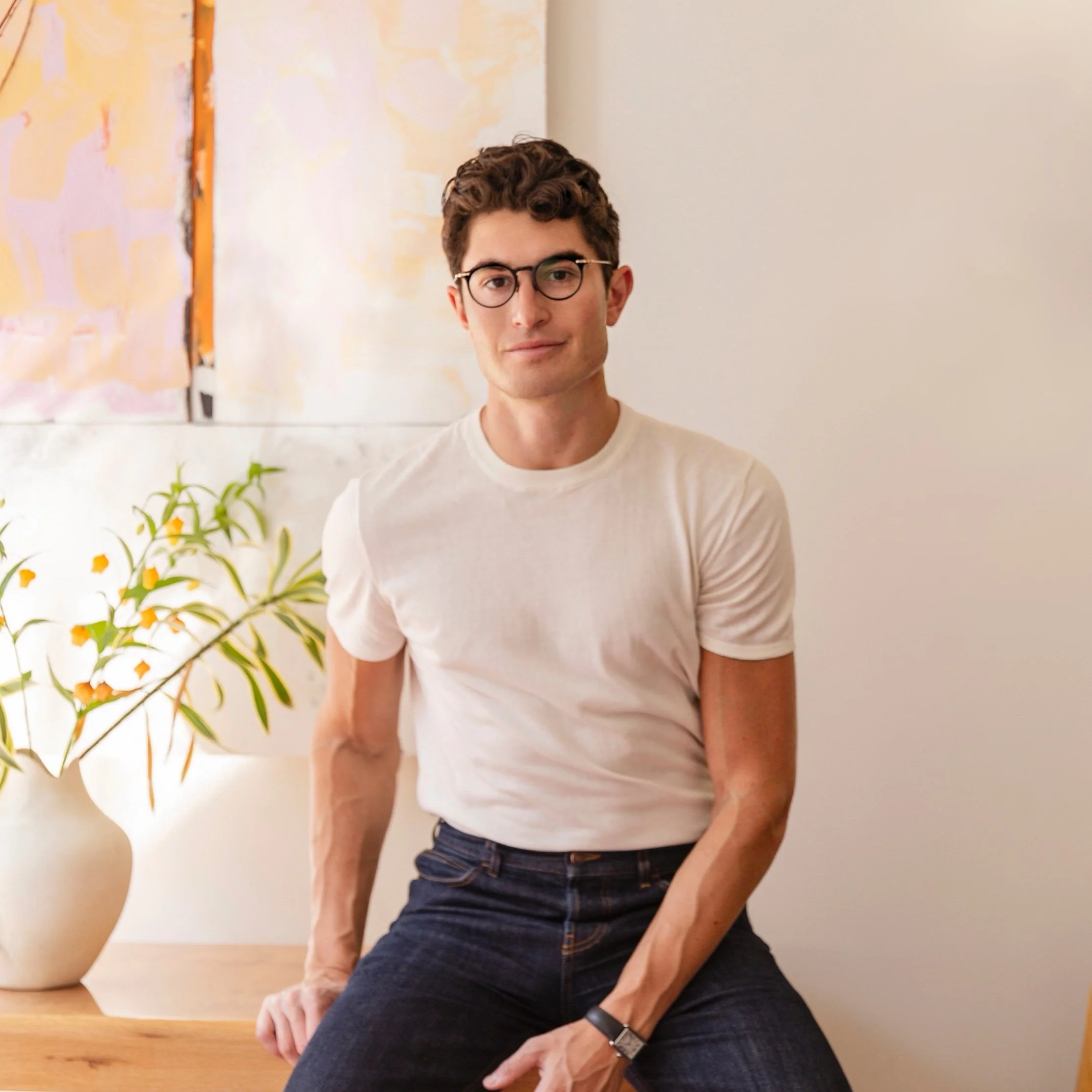 Headshot of Laurence Milstein. Headshot of Larry Milstein. A young man with curly brown hair, wearing glasses and a white t-shirt, sitting against a light-colored wall with a potted plant with orange flowers to his left.