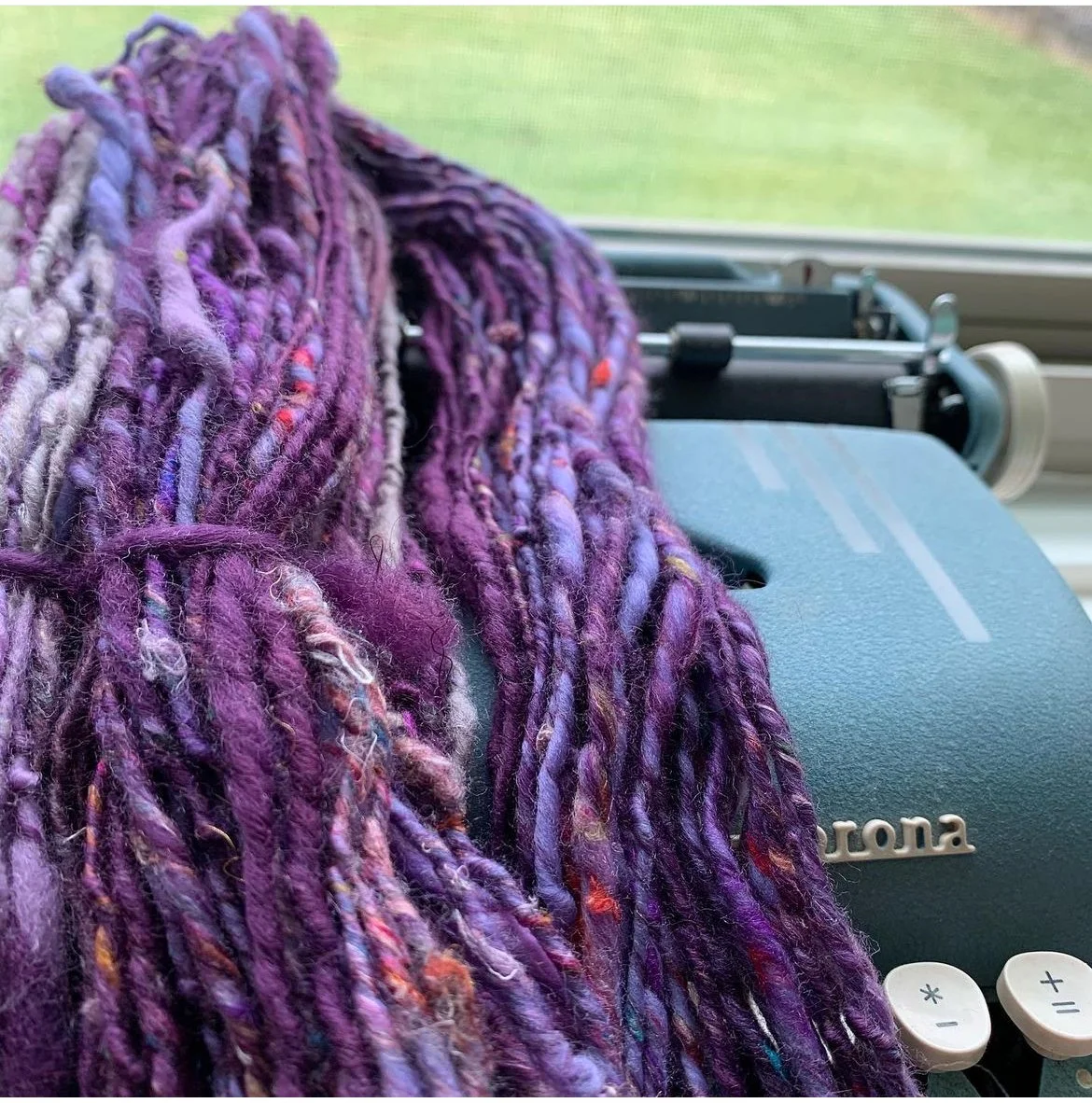 Close-up of purple, multicolored yarn resting on a teal typewriter with a window and green outdoor scene in background.