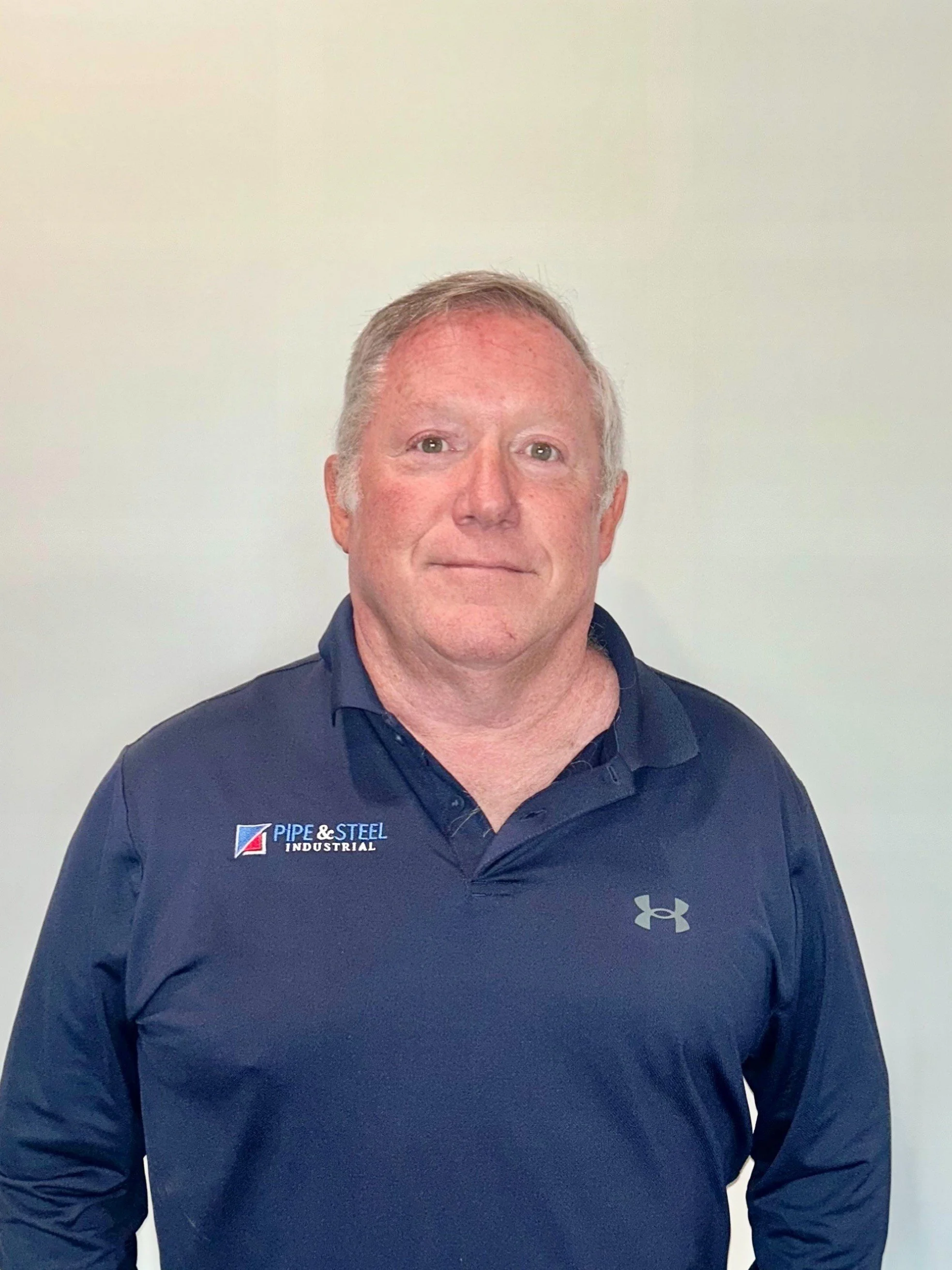 A smiling middle-aged man with short gray hair wearing a navy blue polo shirt with the logos 'Pipe & Steel Industrial' and Under Armour, standing against a plain light-colored background.