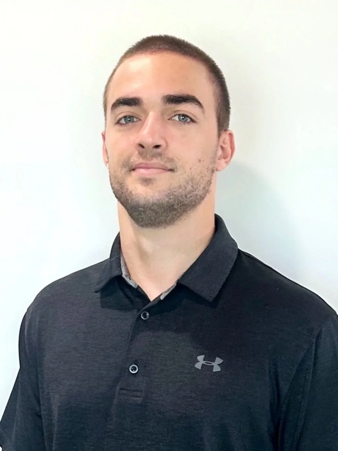 A young man with short brown hair and a beard, wearing a black Under Armour polo shirt, standing in front of a plain white wall.