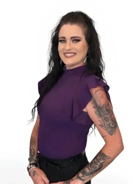 A woman with shoulder-length light brown hair, wearing a black top, silver earrings, and a silver necklace, smiling in front of a plain light background.