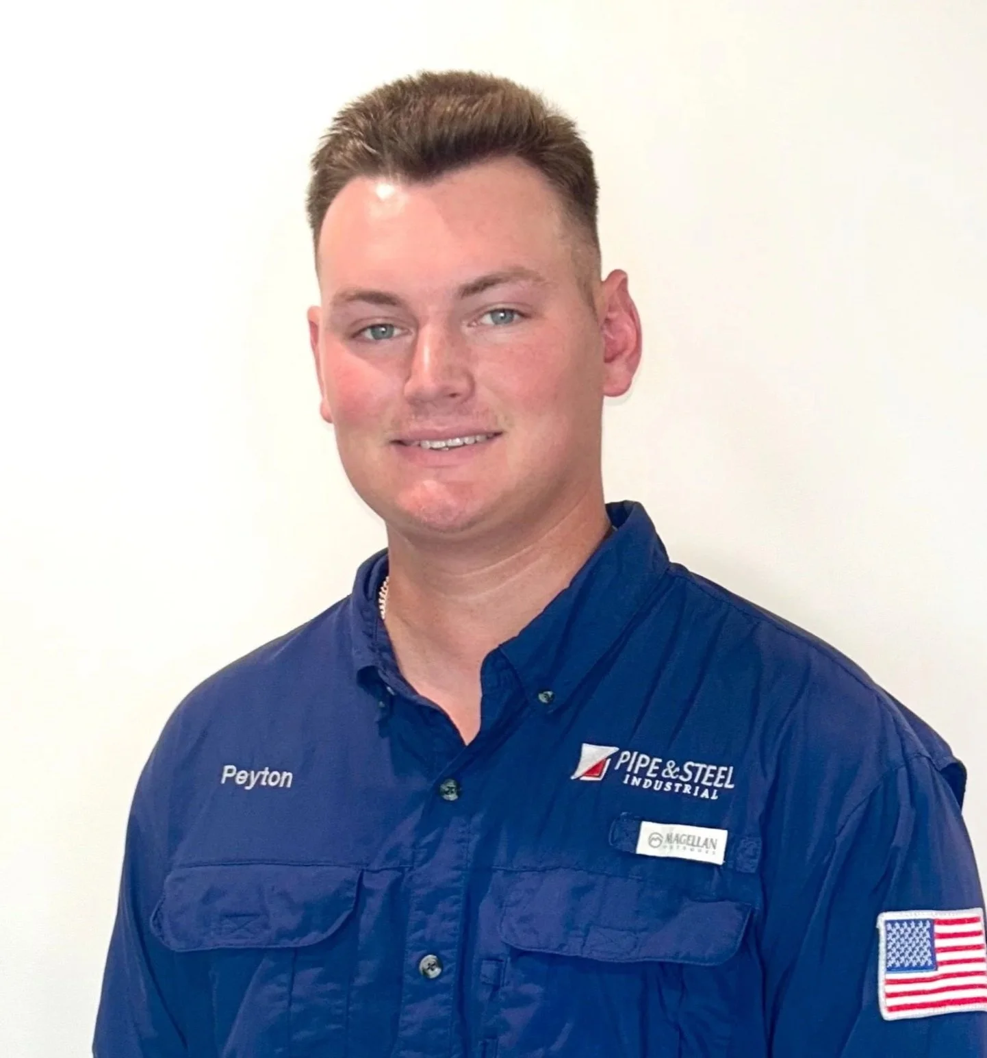 Young man wearing a blue work uniform with patches and name tag, standing against a plain white background.