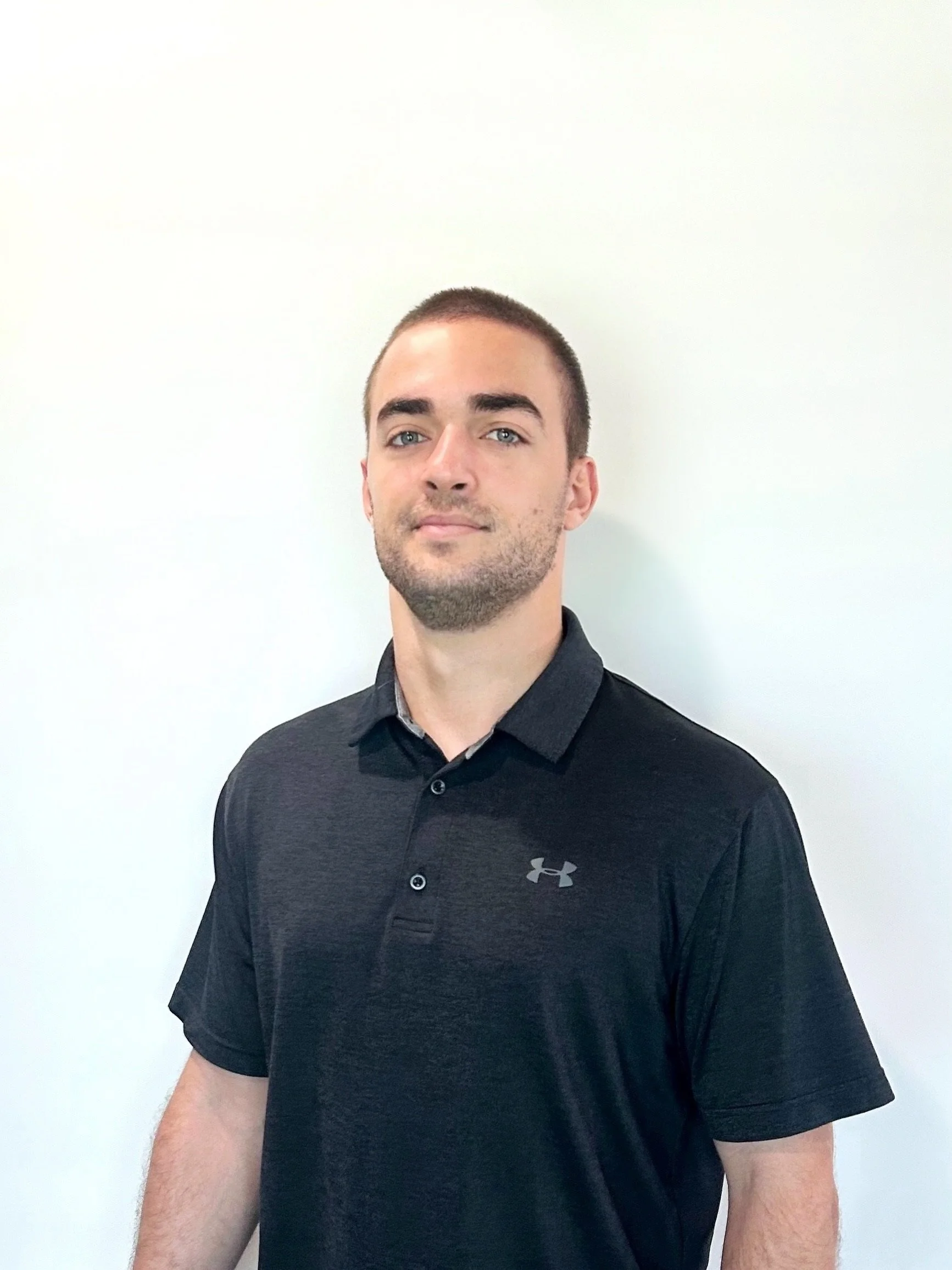 A young man with short brown hair and a beard, wearing a black Under Armour polo shirt, standing in front of a plain white wall.