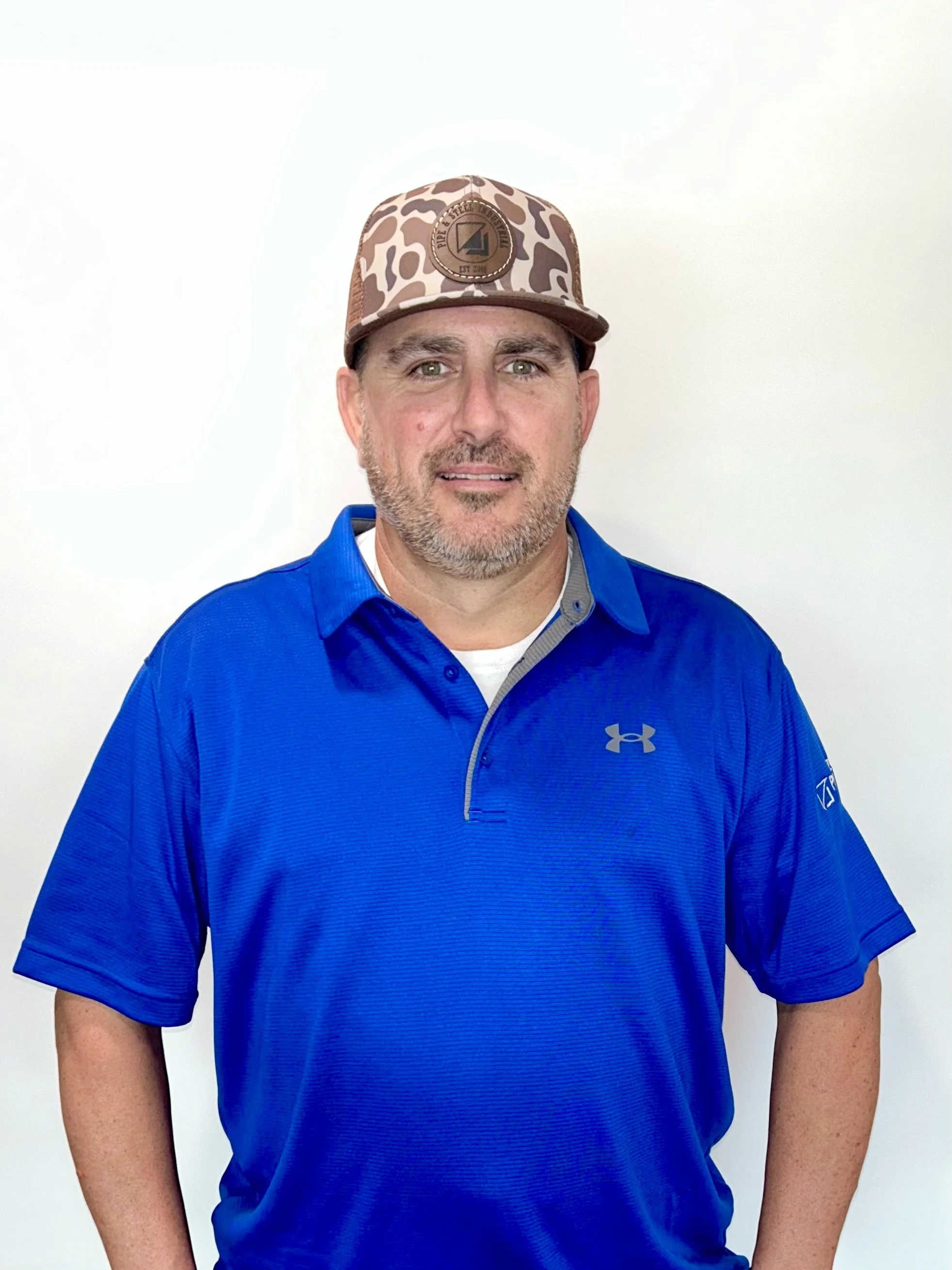 A man wearing a blue Under Armour polo shirt and a leopard-print cap, standing against a plain white background.