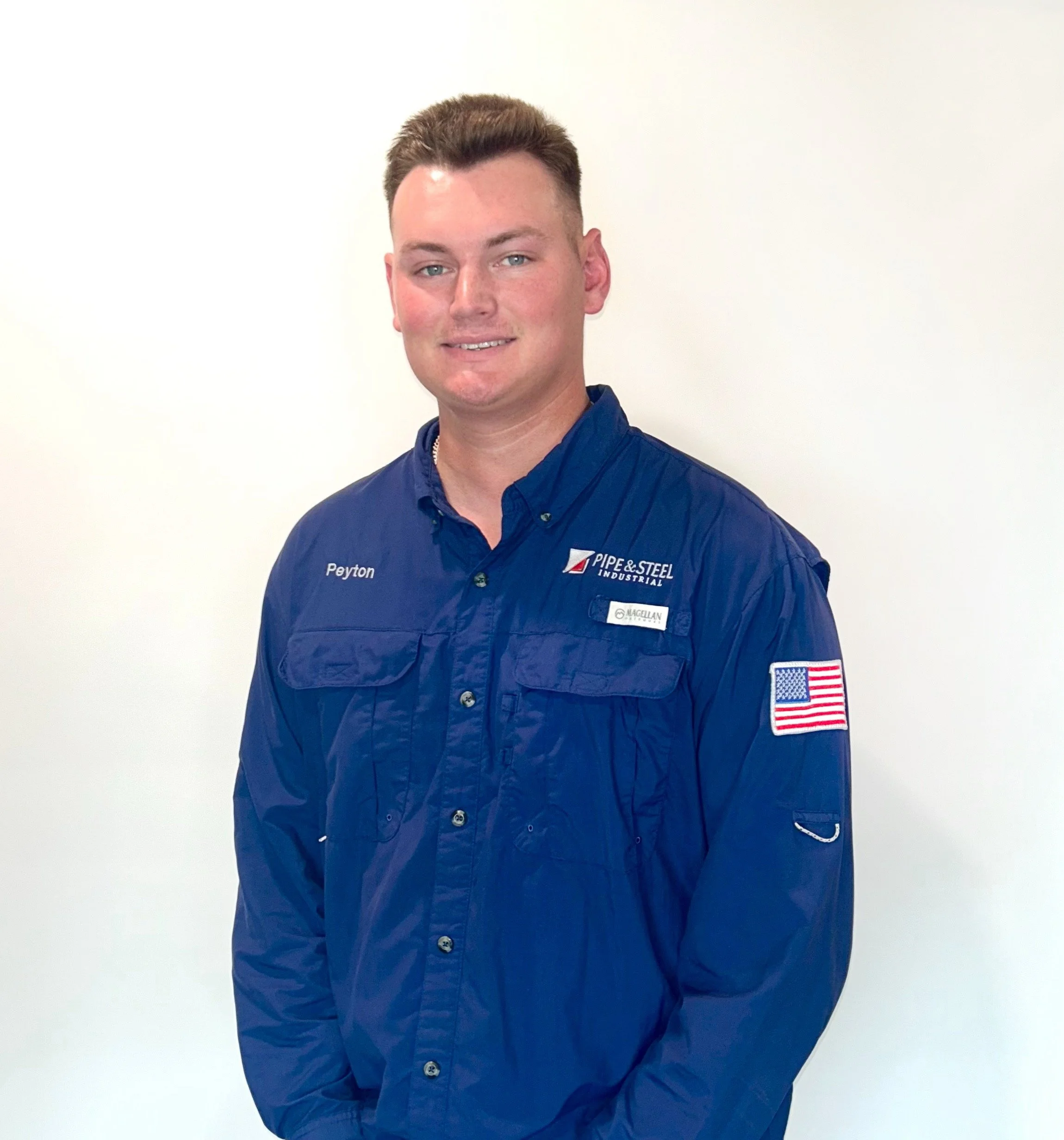Young man wearing a blue work uniform with patches and name tag, standing against a plain white background.