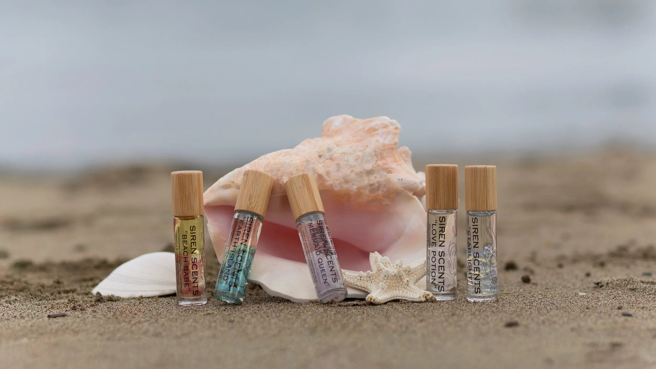 Assorted Siren Scents perfume bottles with gold caps on sandy beach, decorated with a seashell, starfish, and a large conch shell, with ocean waves in background.