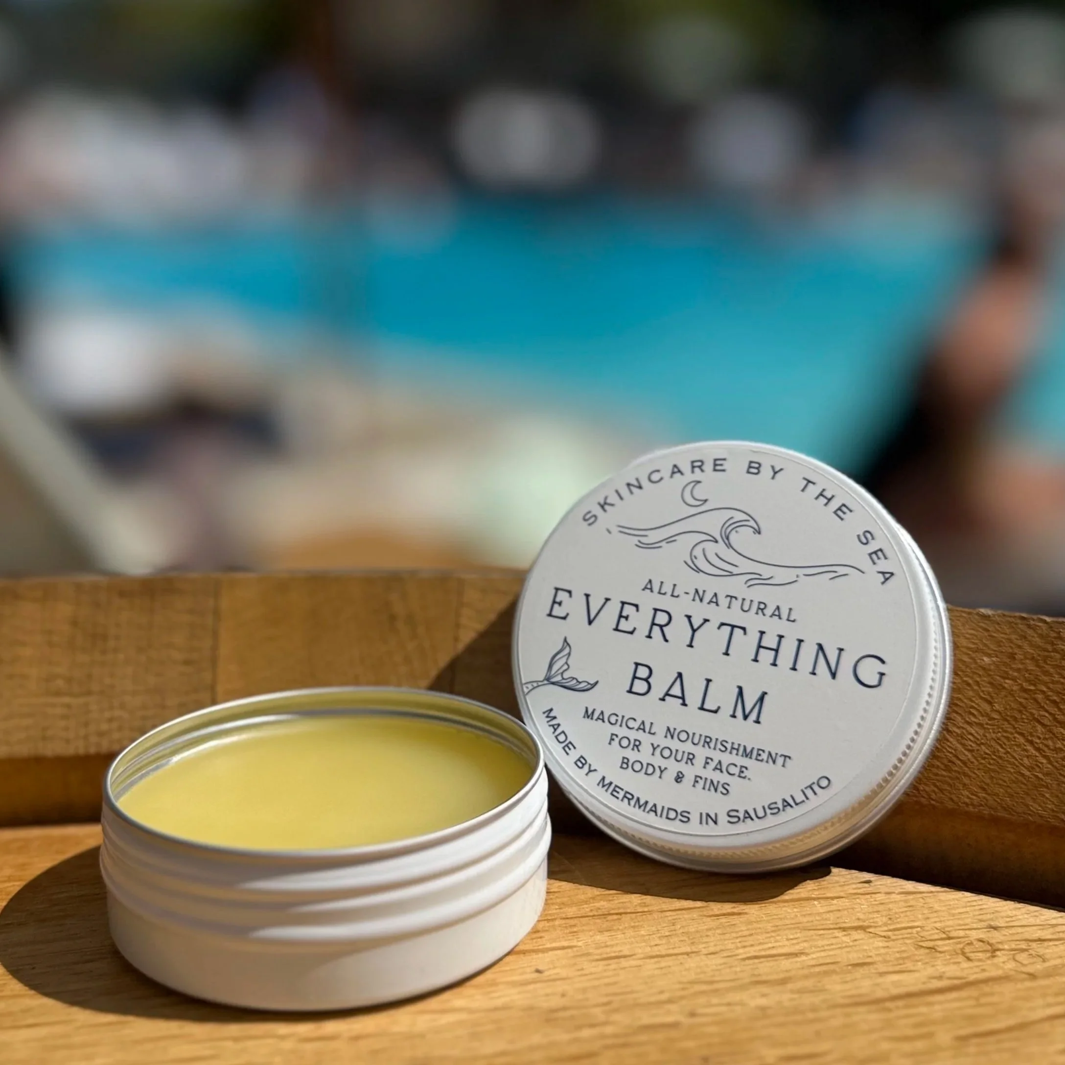Open container of natural all-purpose balm on a rock near the water with a bamboo spatula inside, labeled 'Skincare by the Sea Everything Balm'.