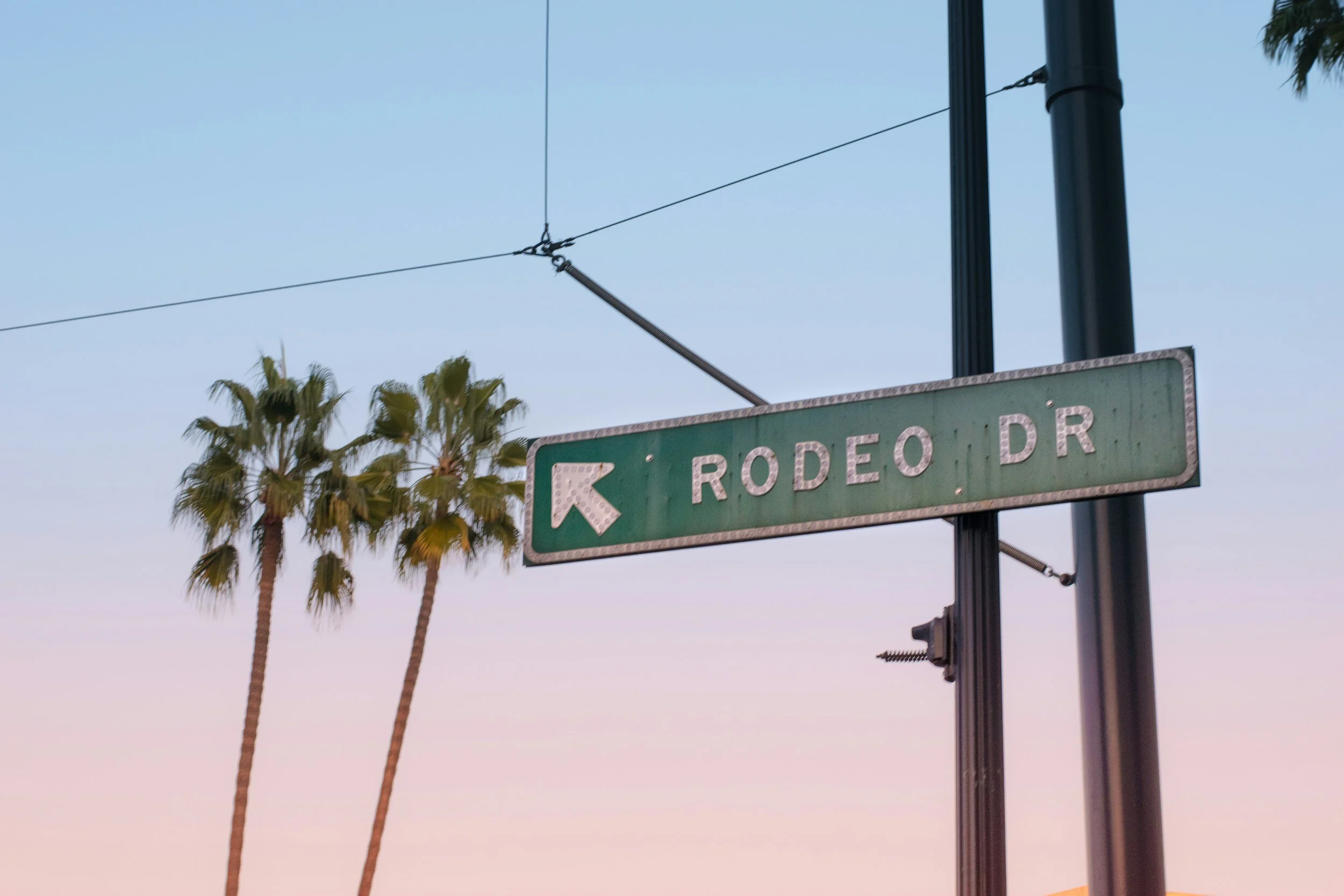 Rodeo Drive street sign with palm trees and a sunset sky background.