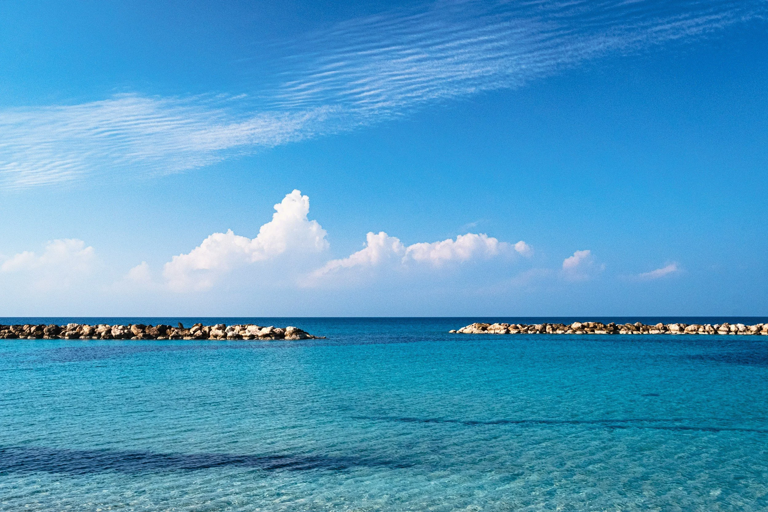 Clear blue ocean with a stone jetty under a bright sky with thin clouds and cumulus clouds on the horizon.