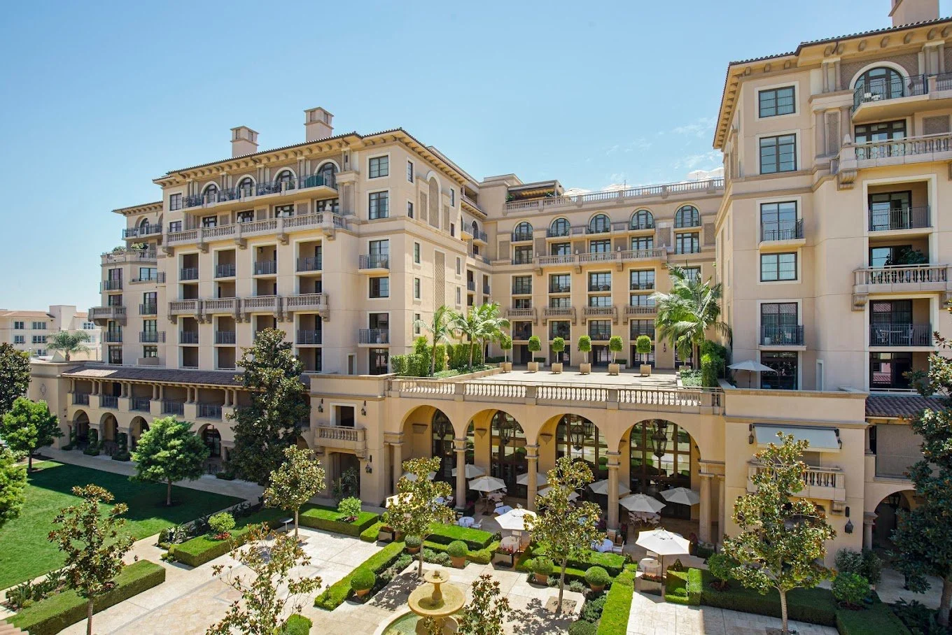 Luxurious hotel building with balconies, arched windows, and a landscaped courtyard featuring a fountain, trees, and outdoor seating with umbrellas.
