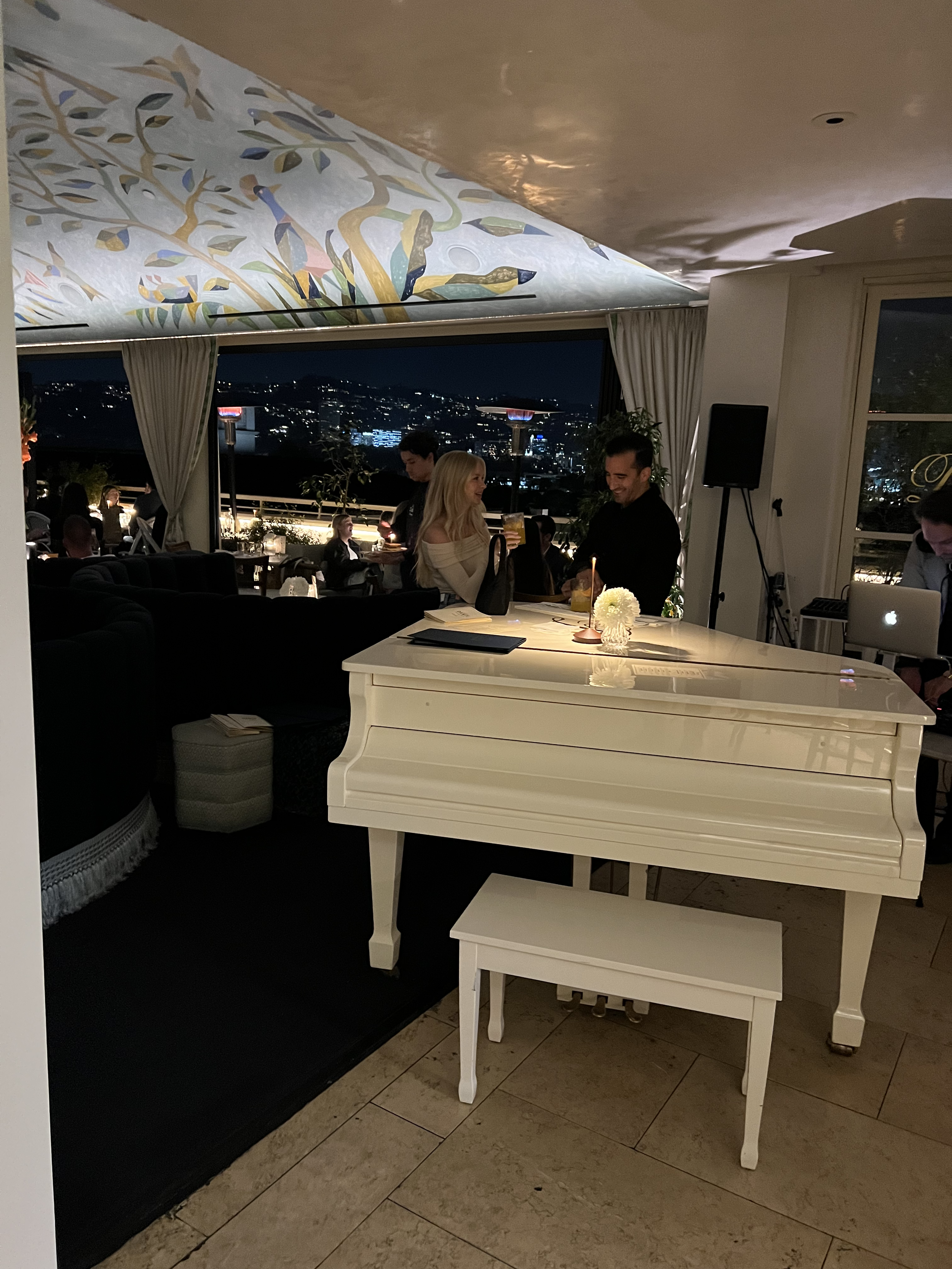 Indoor restaurant at night with a view, featuring a white piano and bench in the foreground, people socializing, and a ceiling mural with colorful designs.