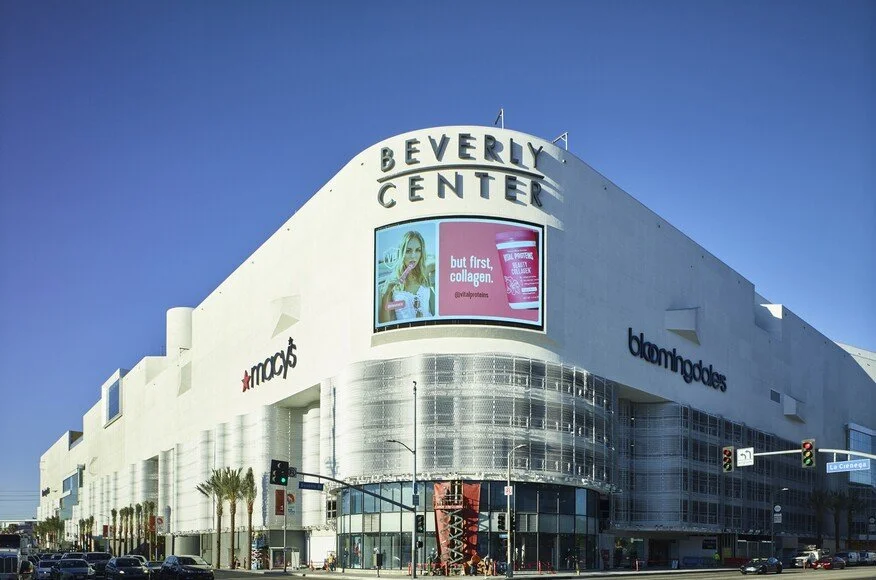Exterior of Beverly Center mall with large Macy's and Bloomingdale's signs, featuring digital advertisement, blue sky background.