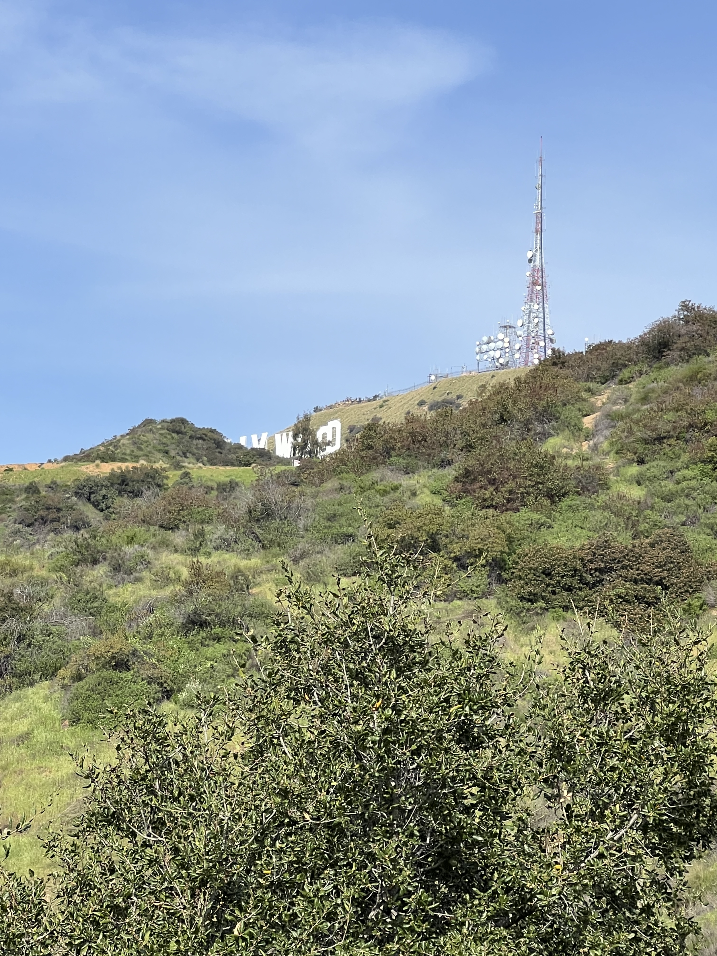 Landscape view of the Hollywood Sign partially obscured by green hills and vegetation, with a telecommunications tower visible on the right under a clear blue sky.