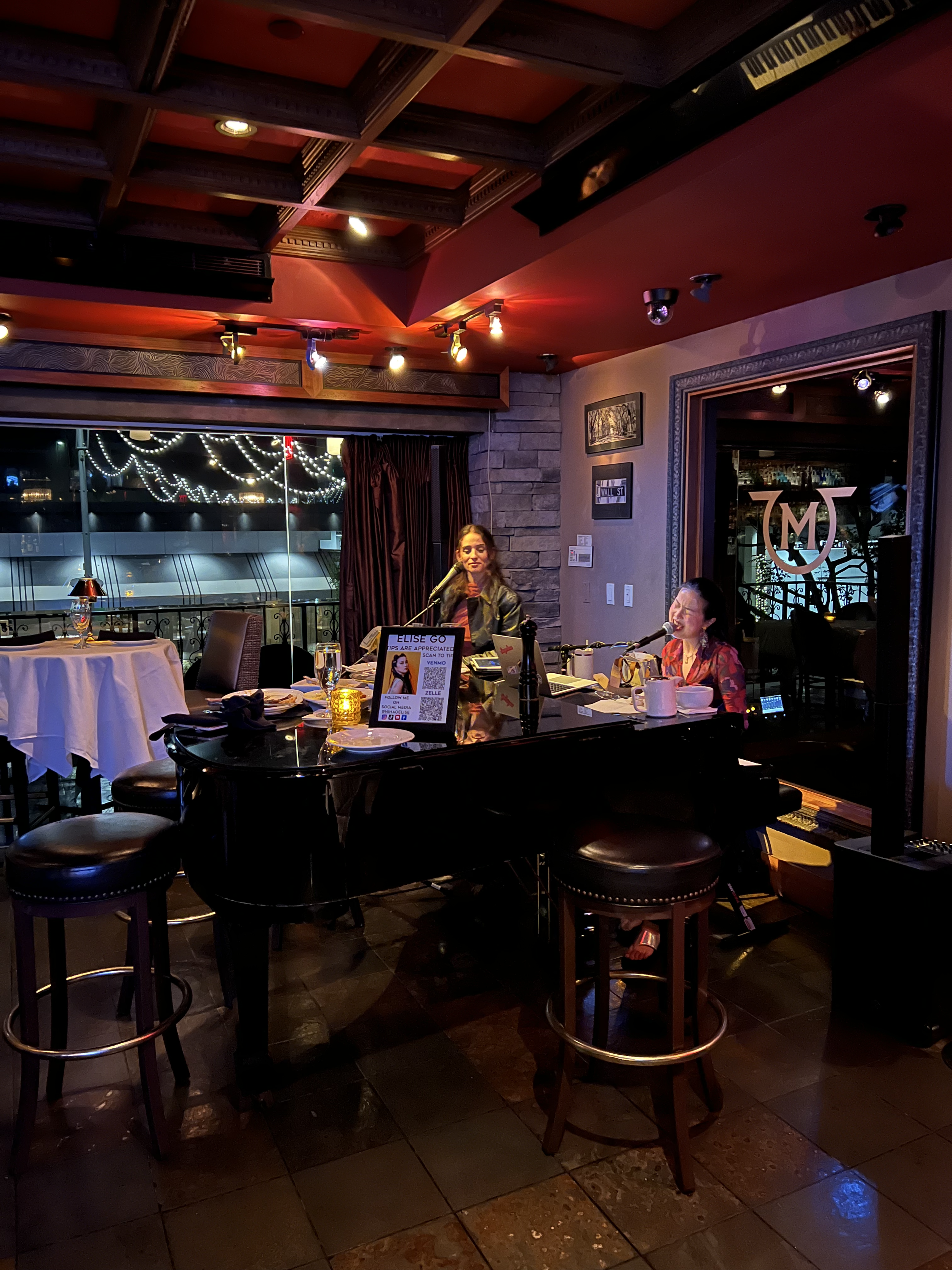 Two people performing at a piano inside a dimly lit bar with wooden ceiling and decorative lights. A microphone and sheet music are on the piano. Tables with white tablecloths and chairs are visible around.