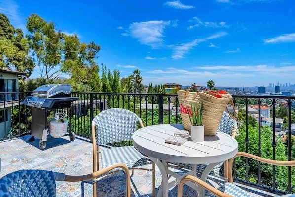Outdoor patio with a white round table and blue chairs, decorative floor tiles, and a barbecue grill. A potted plant and a decorative item sit on the table. Cityscape and trees visible in the background under a blue sky.