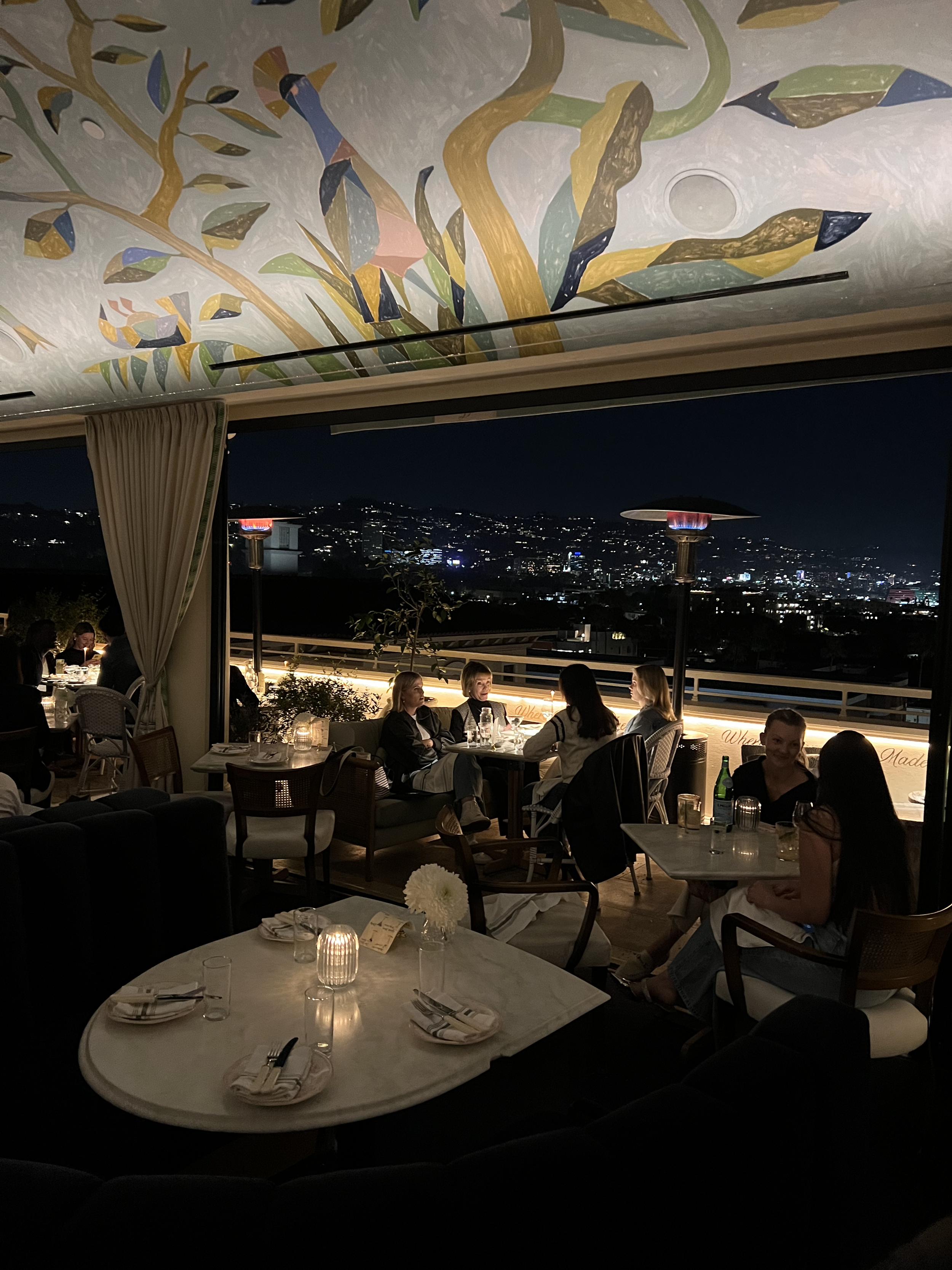 Outdoor rooftop restaurant at night with city lights view, featuring diners seated at tables under artistic ceiling decor.