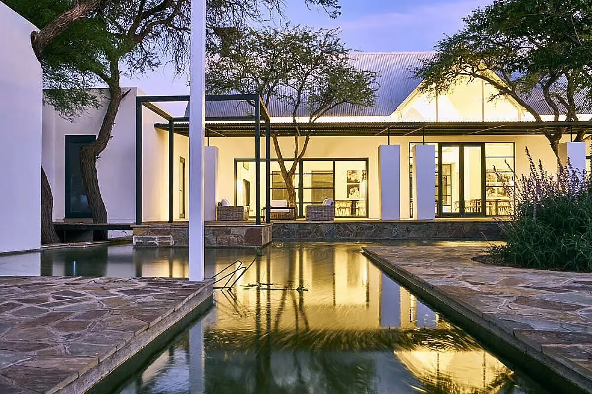 Modern house with large windows, outdoor patio, and reflecting pool surrounded by trees.