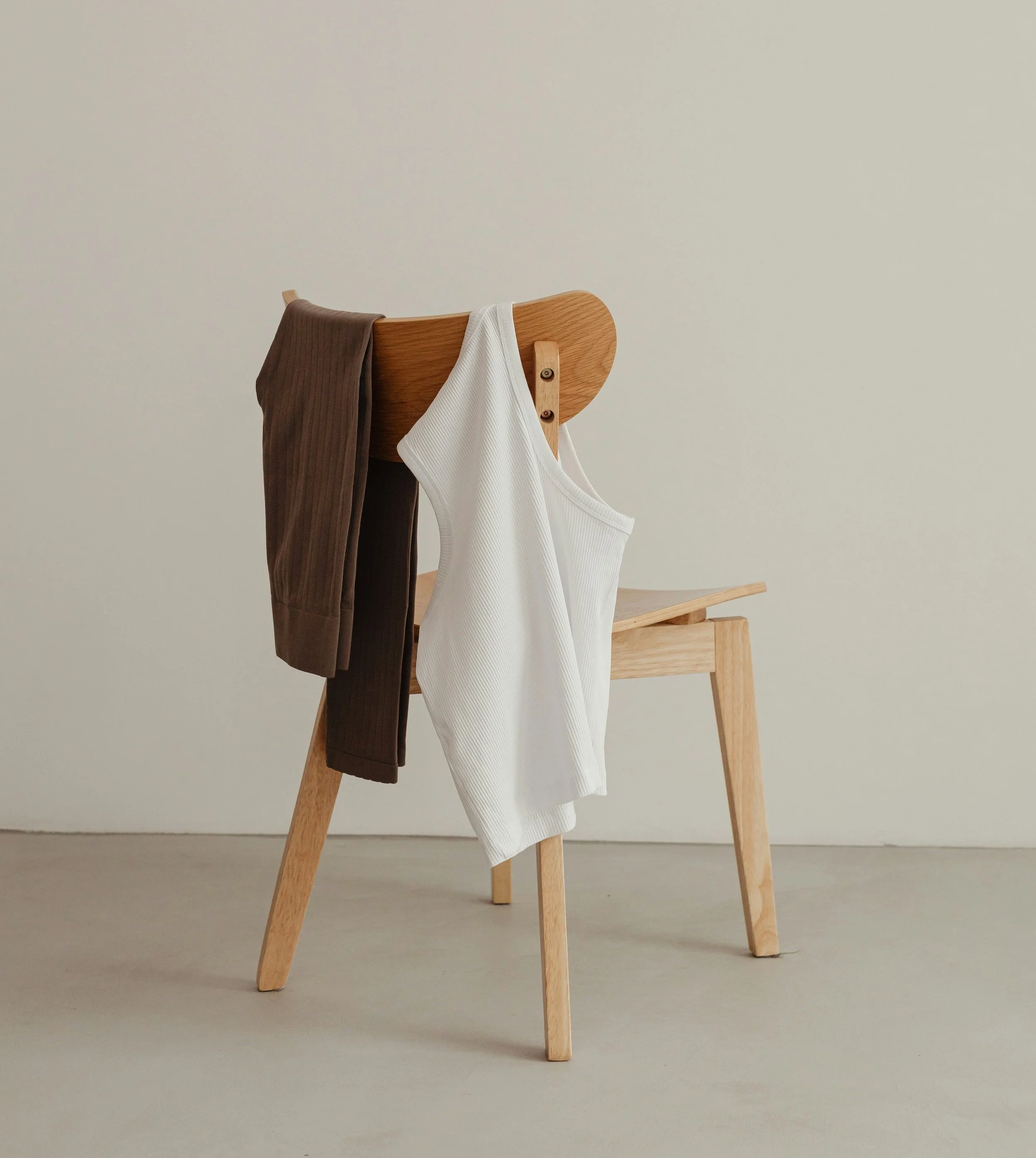 Wooden chair with white tank top and brown pants draped over it against a plain background.