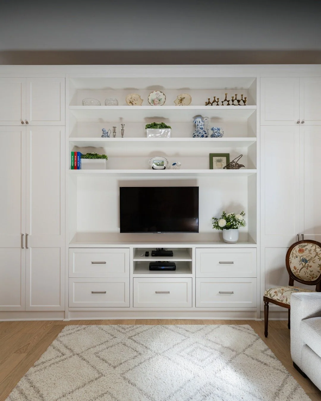 This custom living room built-in creates a seamless home for the TV while balancing storage and style. From porcelain accents to greenery and art, every shelf tells a story without the clutter. Paired with traditional touches like a floral chair, the