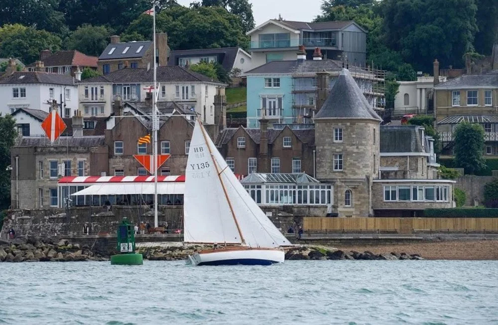 A sailboat near a coastal town with various houses, including a stone castle-like building, under cloudy weather.